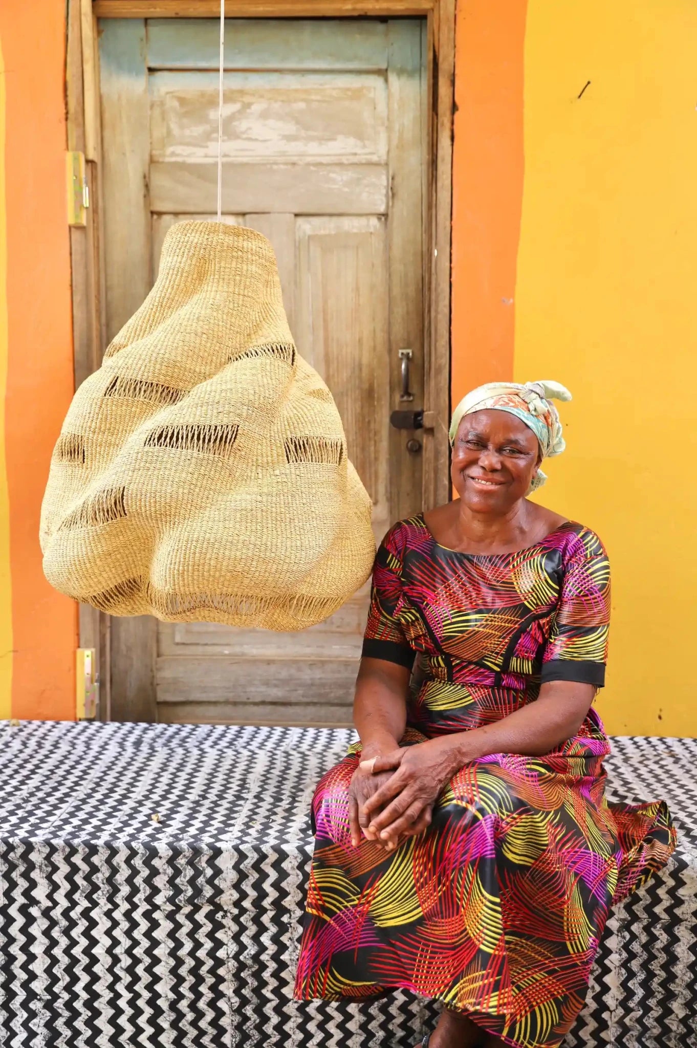 Woman in a colorful dress sitting on a bench with a woven basket hanging above her against a yellow wall.