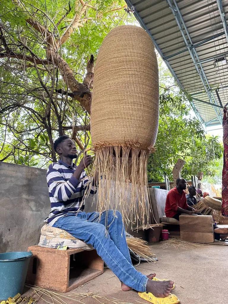 A tall natural straw basket hanging upside down while it is woven