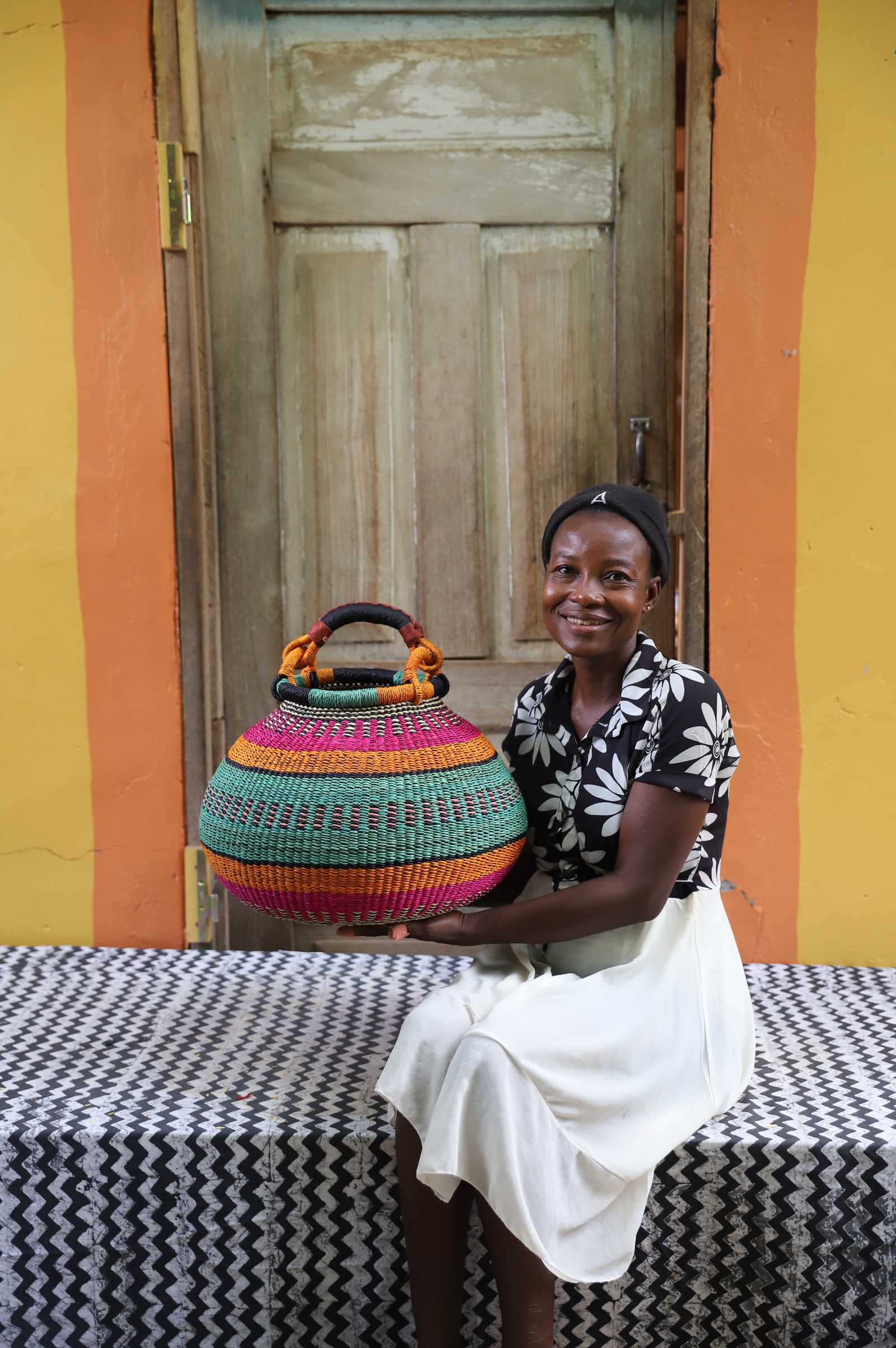 Large handwoven Bolga pot basket in bold bands of pink, teal and amber, held by a smiling woman in Bolgatanga.
