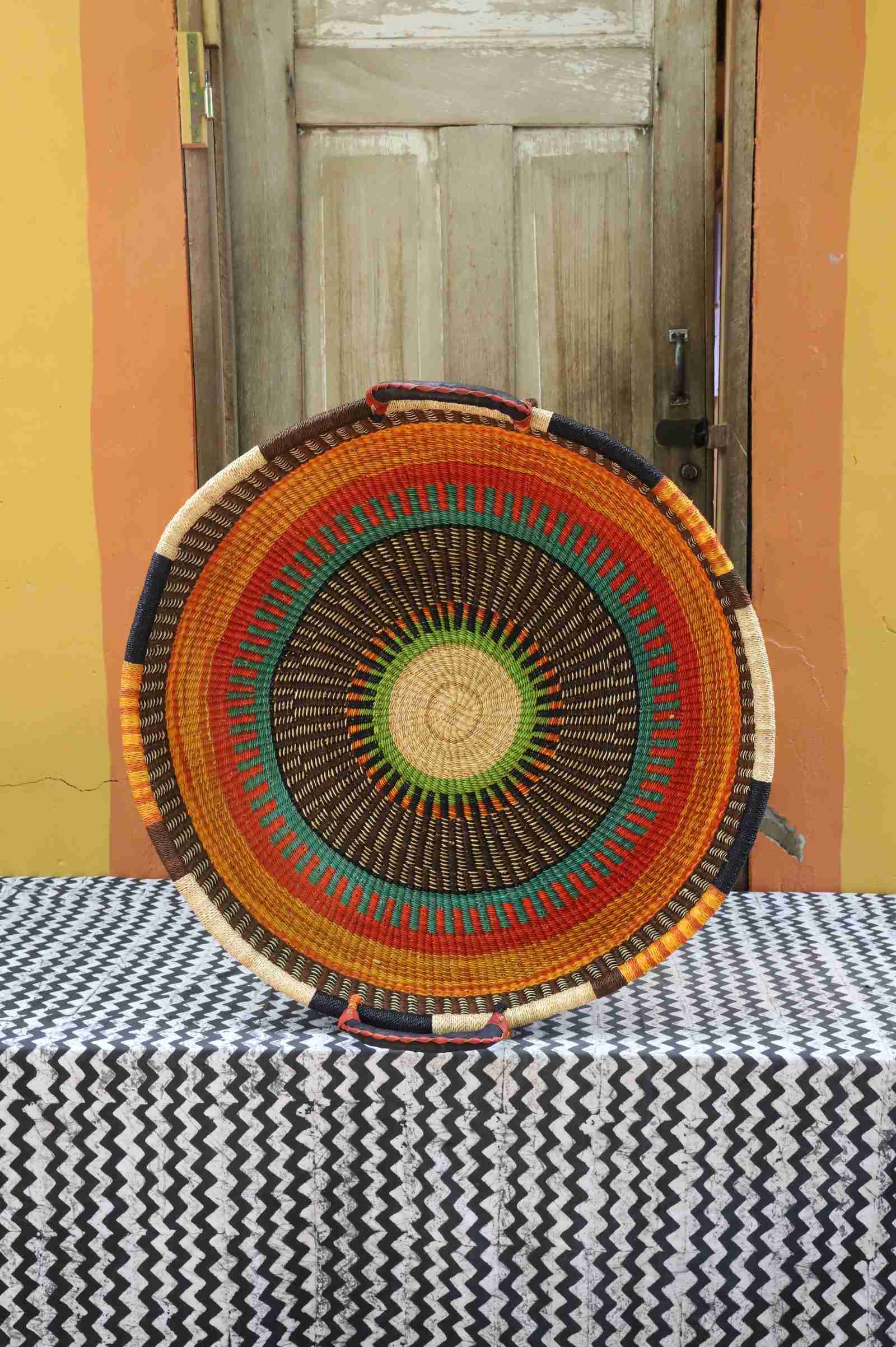 Large handwoven Bolga wall tray disc with orange, teal and green concentric bands and dark radial rays.