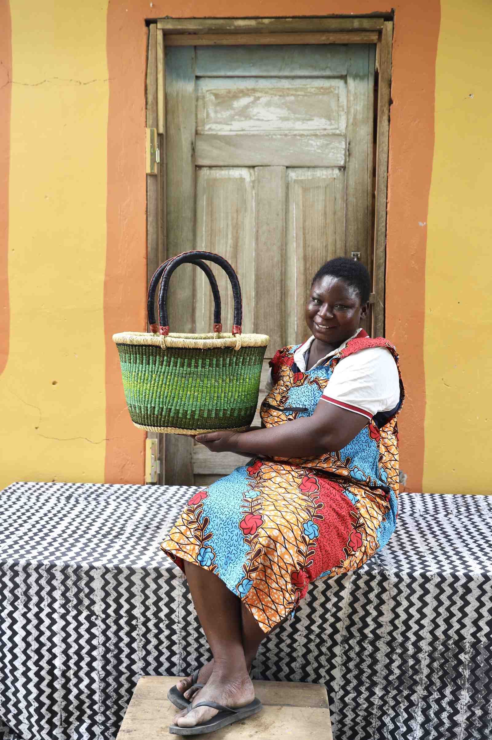 Small oval Bolga shopping basket in natural with green bands, held by a seated woman in red.