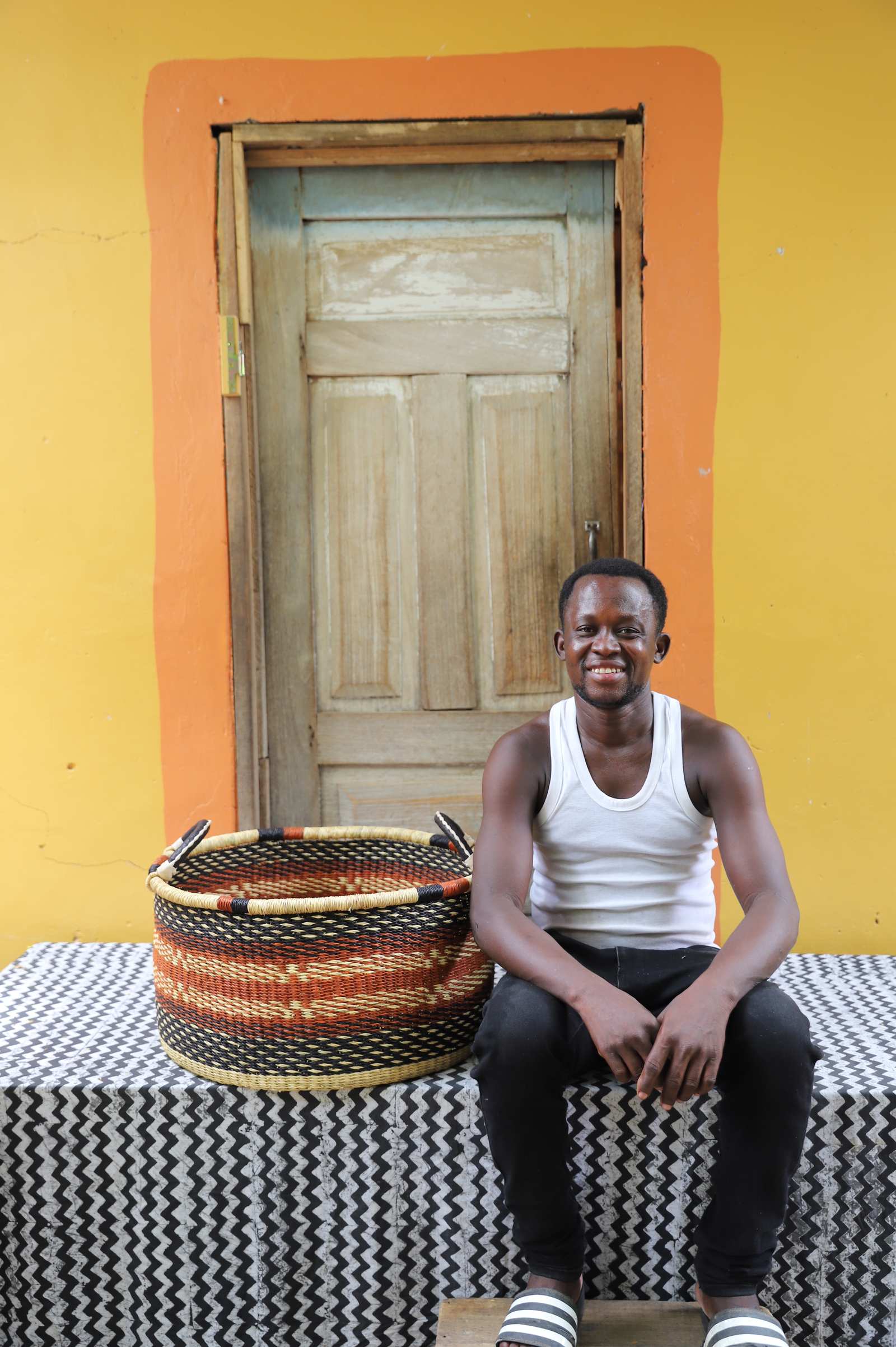 Large handwoven Bolga storage tub in natural straw with orange and black diamonds, beside a seated man.
