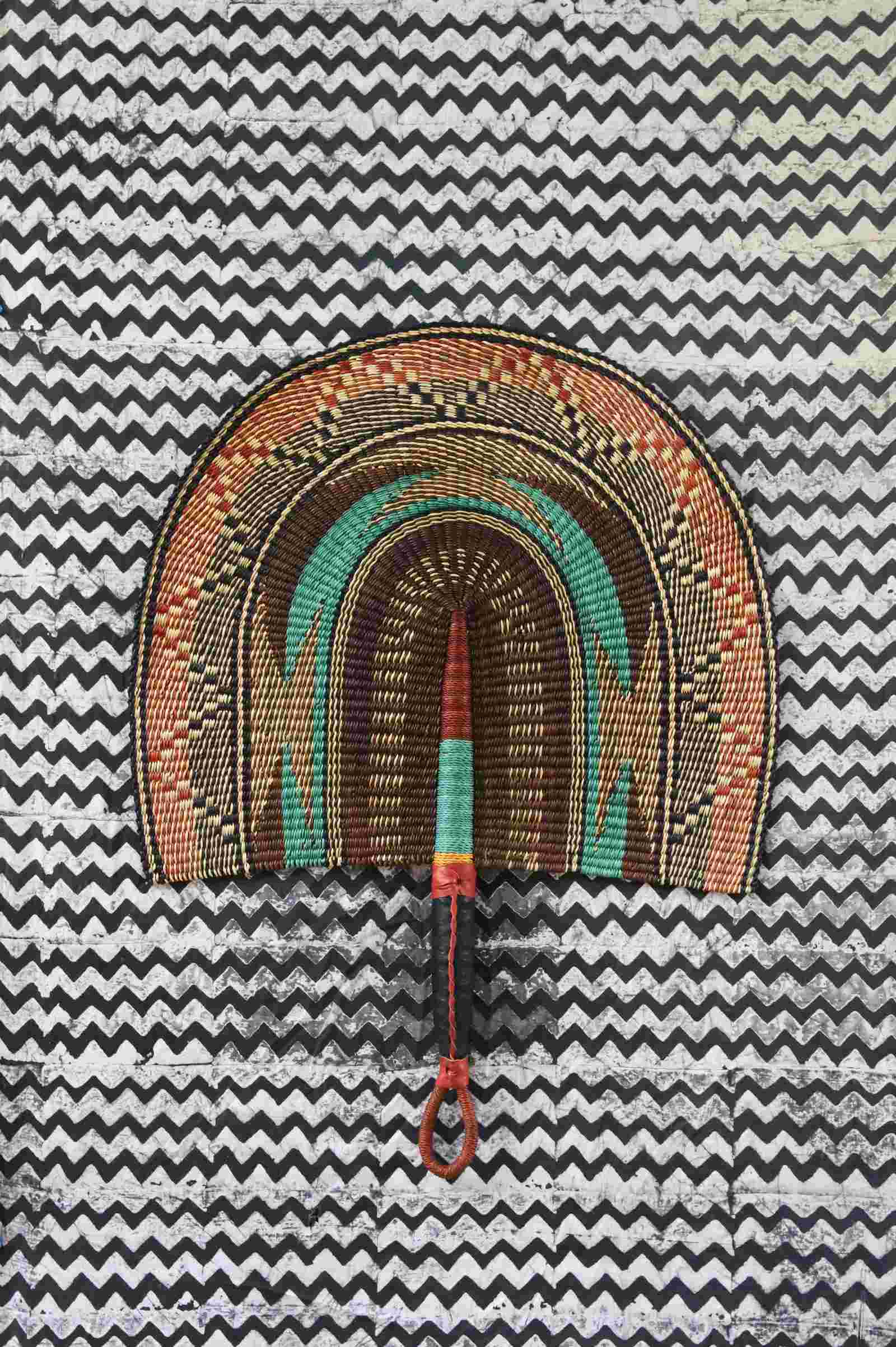 Naba Penga wall fan in natural straw with a teal arch and orange patterned border, laid on a chevron cloth.