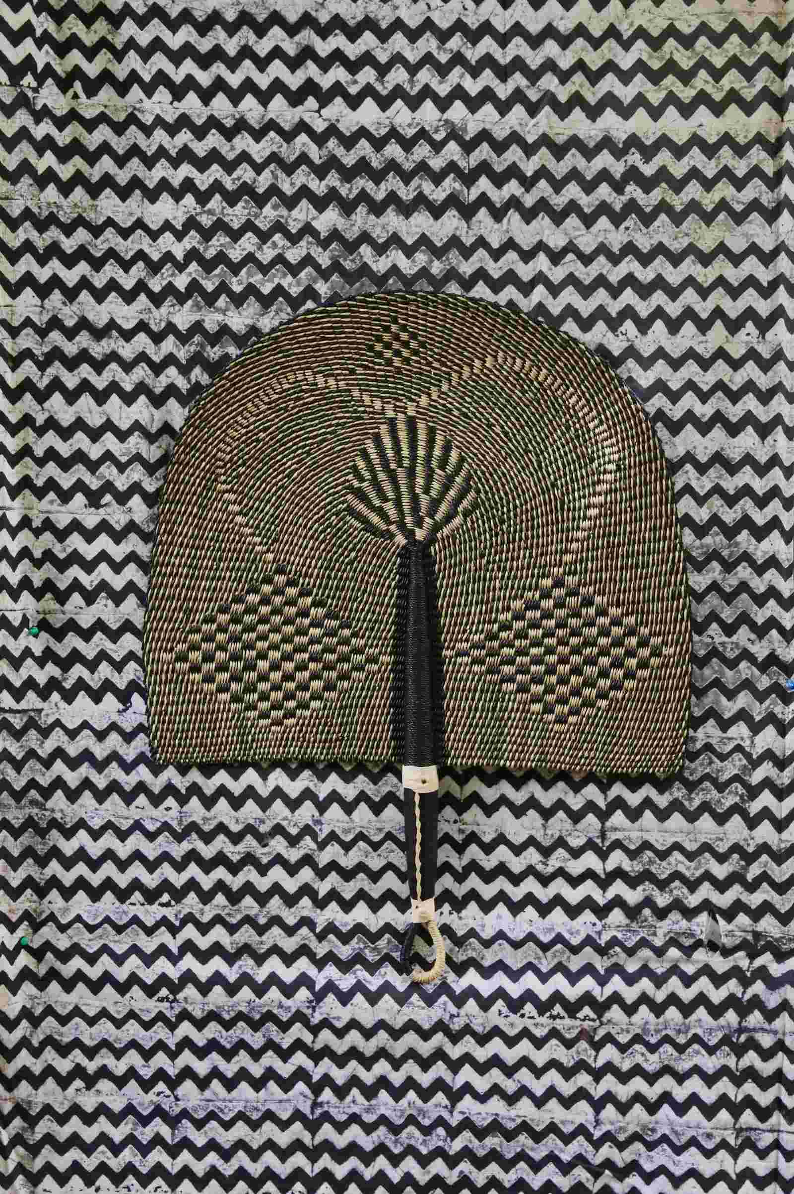 Naba Penga handwoven wall fan in natural straw and black geometric check pattern, on a chevron-print backdrop.