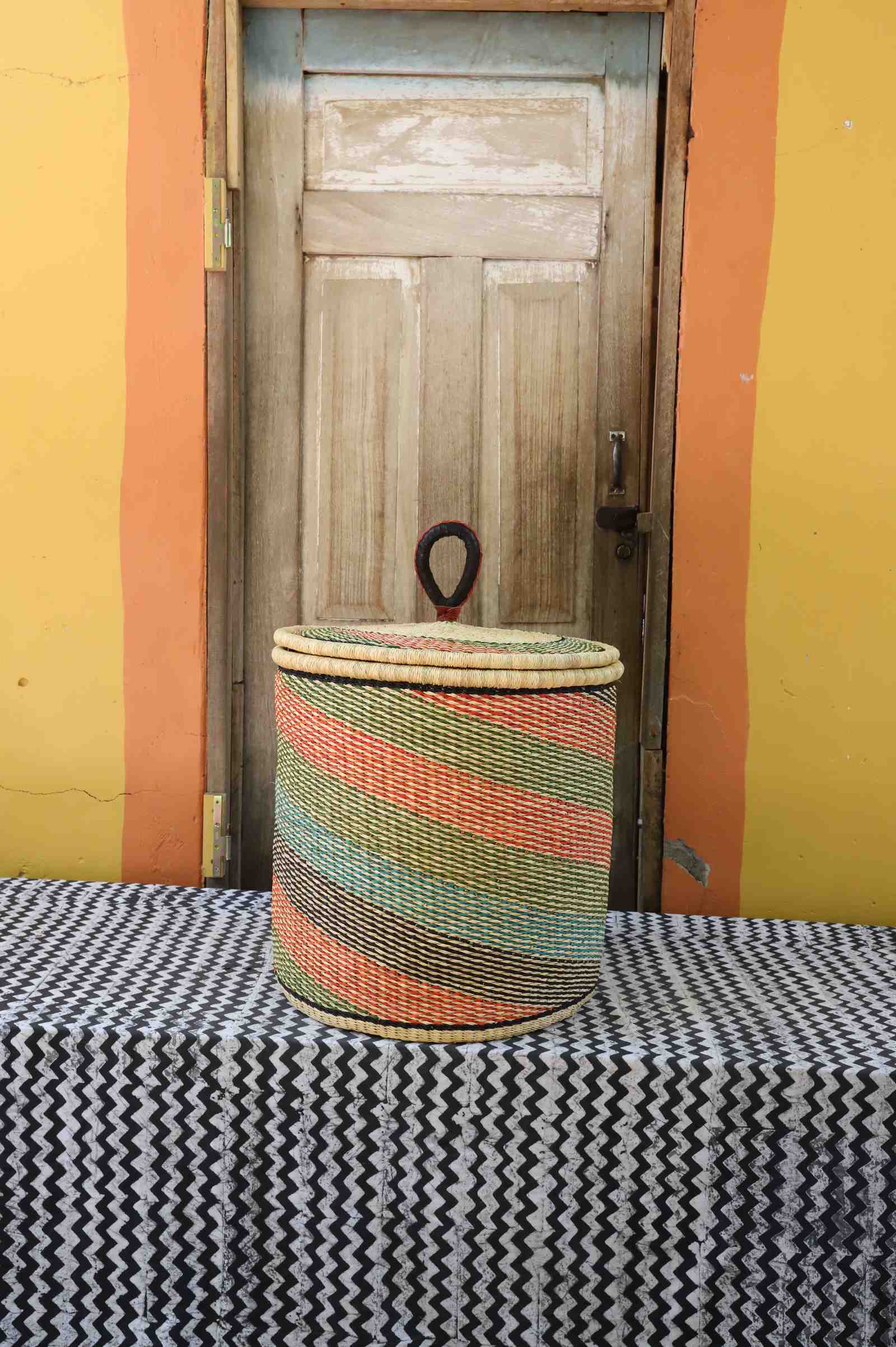 Handwoven Bolga laundry basket with coiled rim and lid in natural straw with fine pink, green and orange diagonal stripes.
