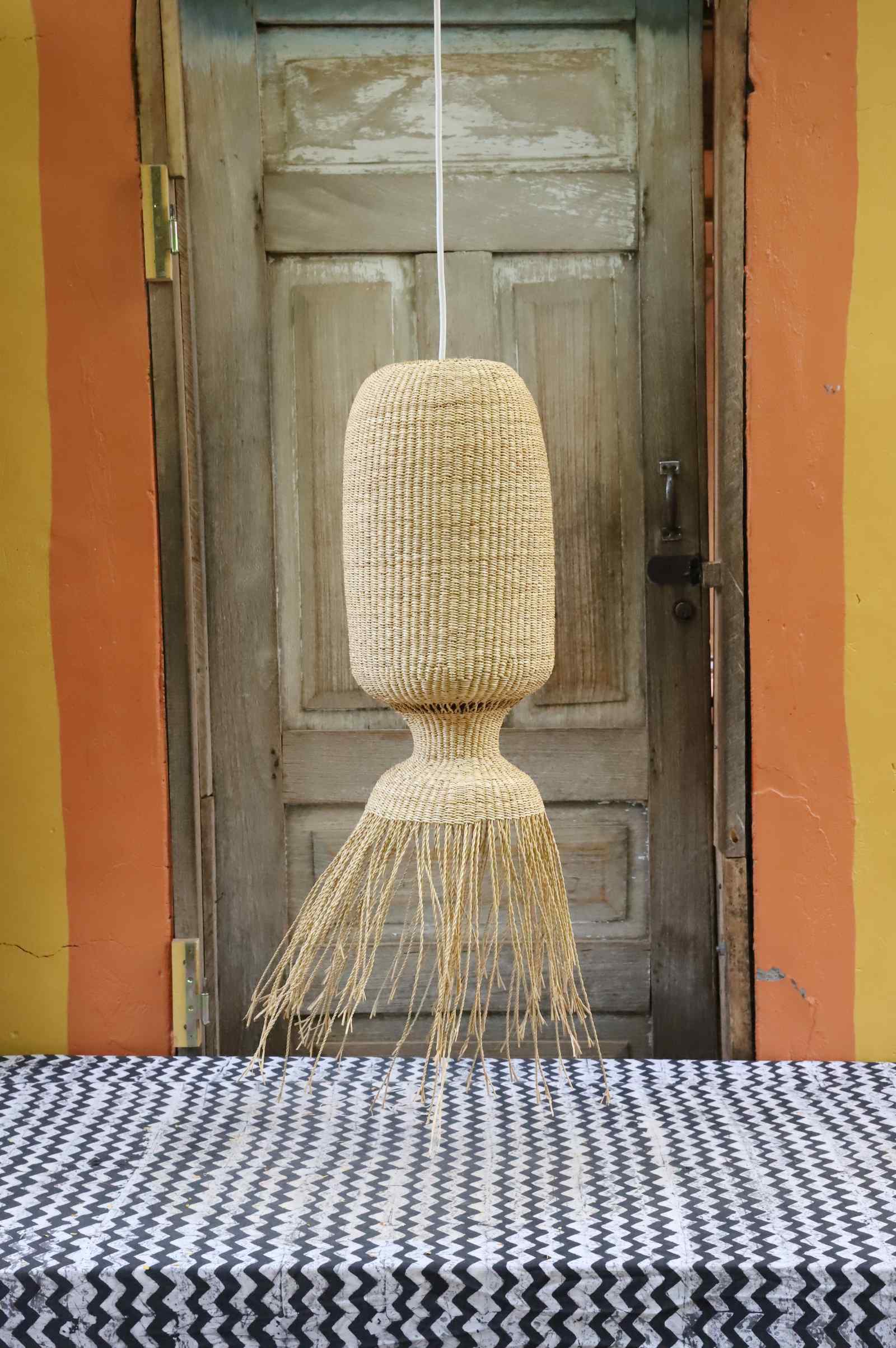 Elephant grass Abyssal pendant lampshade with tapered fringe base, hung against a weathered door.