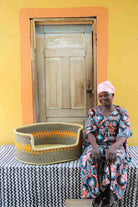 Large handwoven Bolga pet bed in natural straw with fine orange stripes, beside a seated woman in a printed dress.