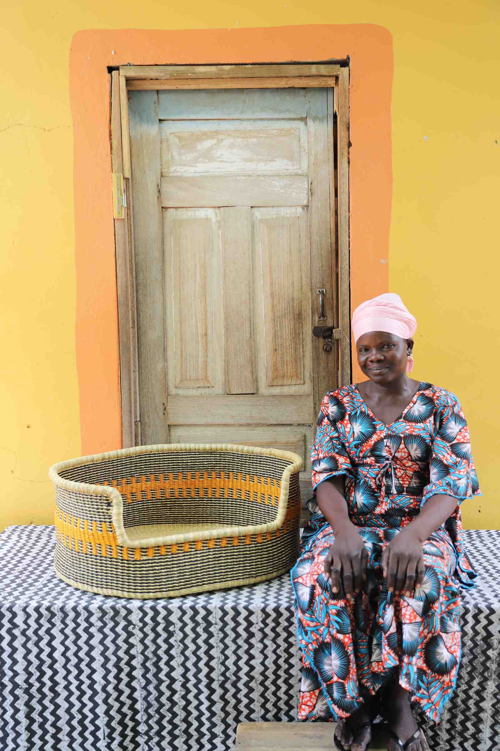 Large handwoven Bolga pet bed in natural straw with fine orange stripes, beside a seated woman in a printed dress.