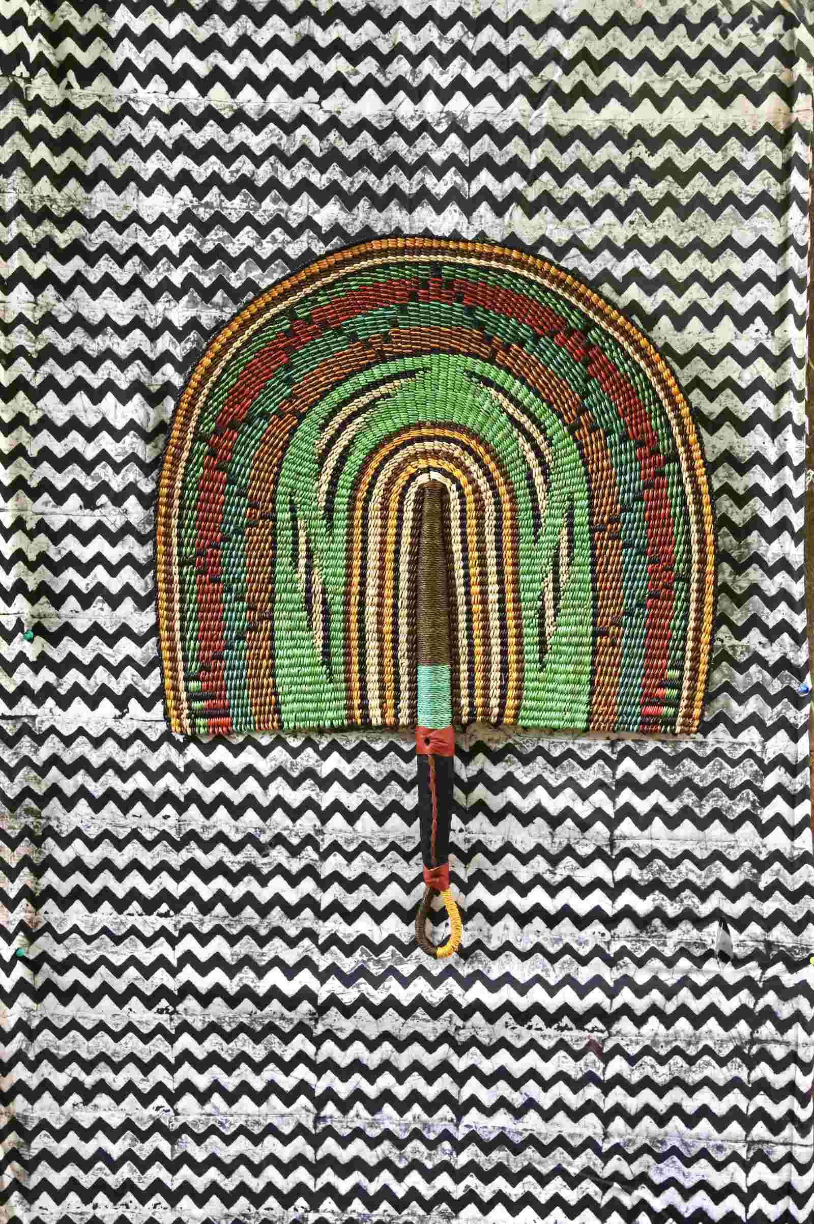 Naba Penga handwoven wall fan with arch bands in green, terracotta and amber, on a chevron-print fabric backdrop.