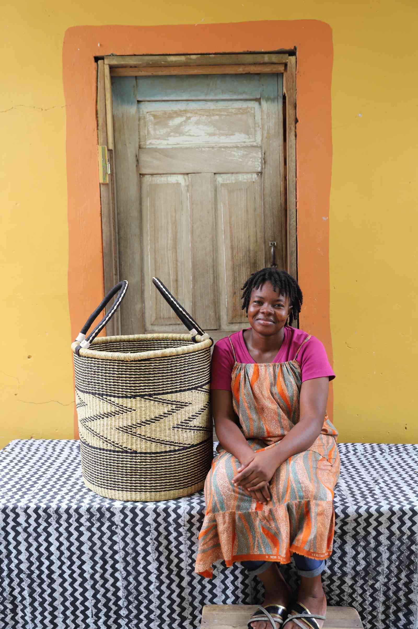 Large handwoven Bolga laundry basket in natural with fine black zigzag wave, beside a seated woman in orange.