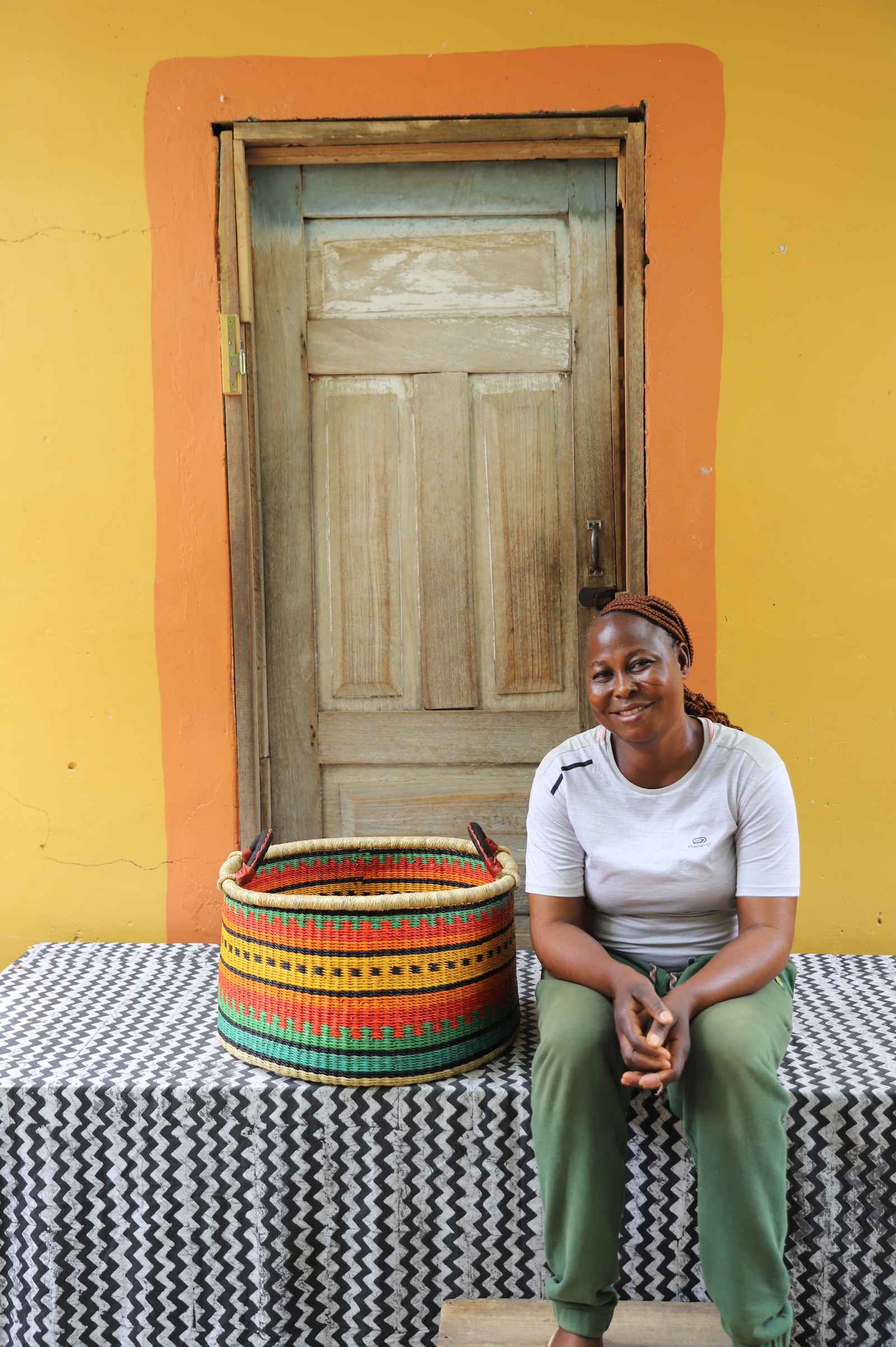 Medium handwoven Bolga storage tub in green with orange and yellow bands, beside a seated woman in white.