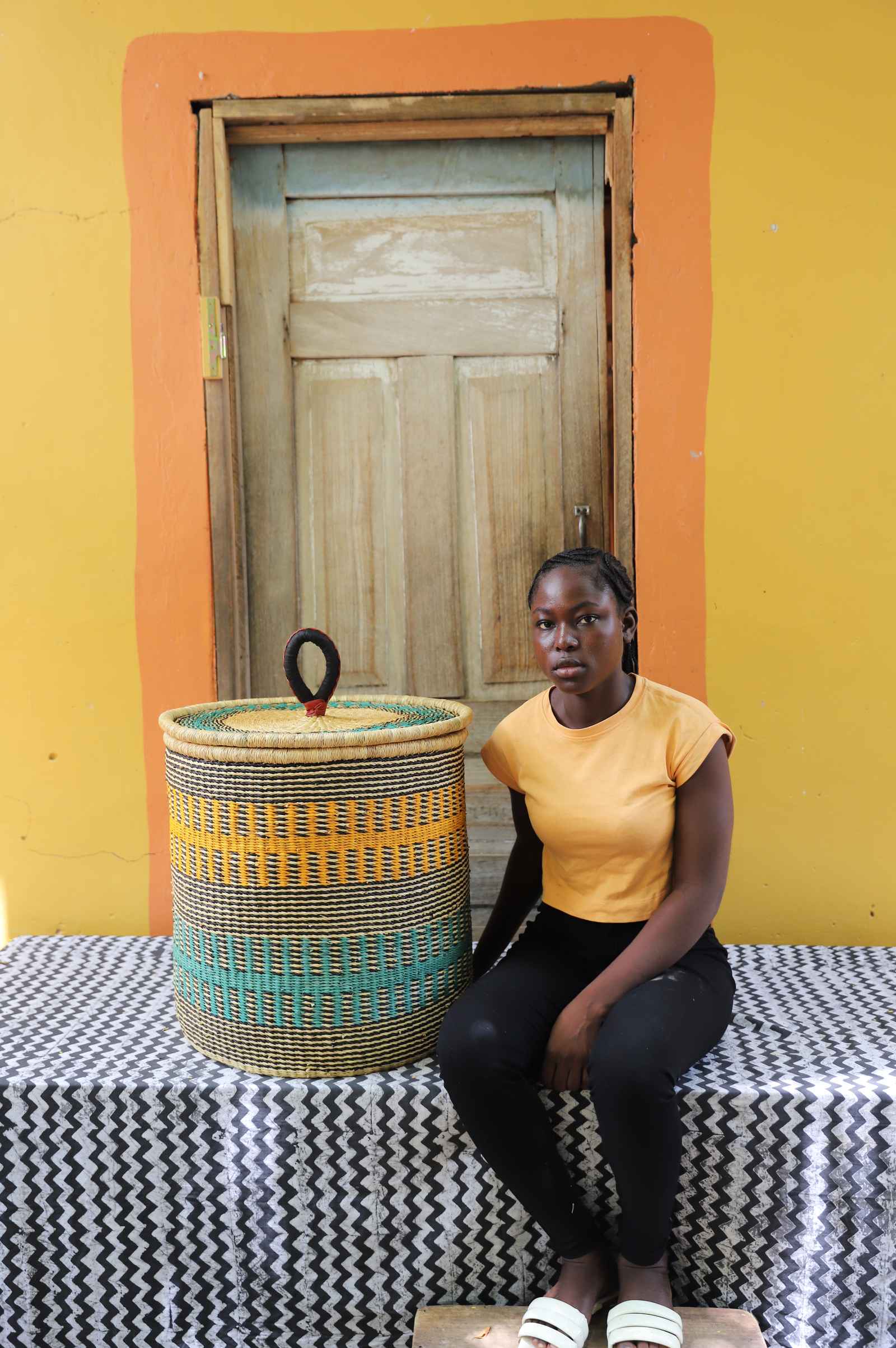 Handwoven Bolga laundry basket in natural straw with yellow and teal bands, beside a seated woman in yellow.