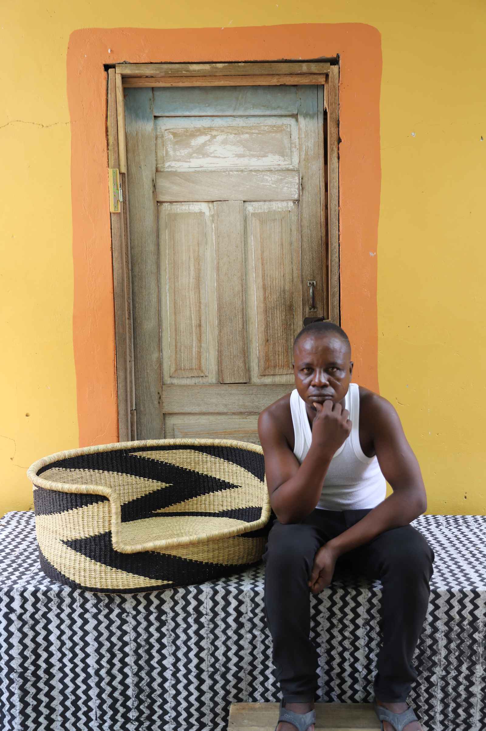 Large handwoven Bolga pet bed in natural straw with bold black chevron, beside a seated man in a white vest.