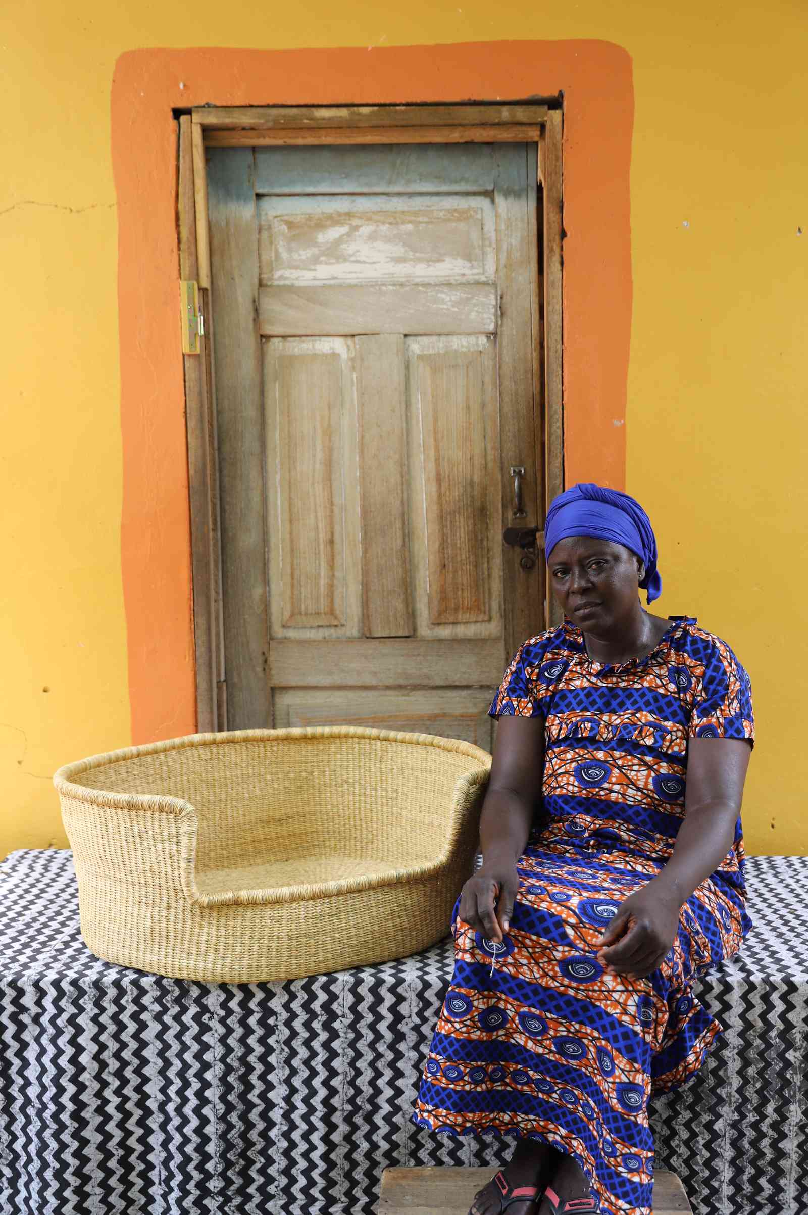 Large handwoven Bolga pet bed in plain natural straw, beside a seated woman in a printed dress.