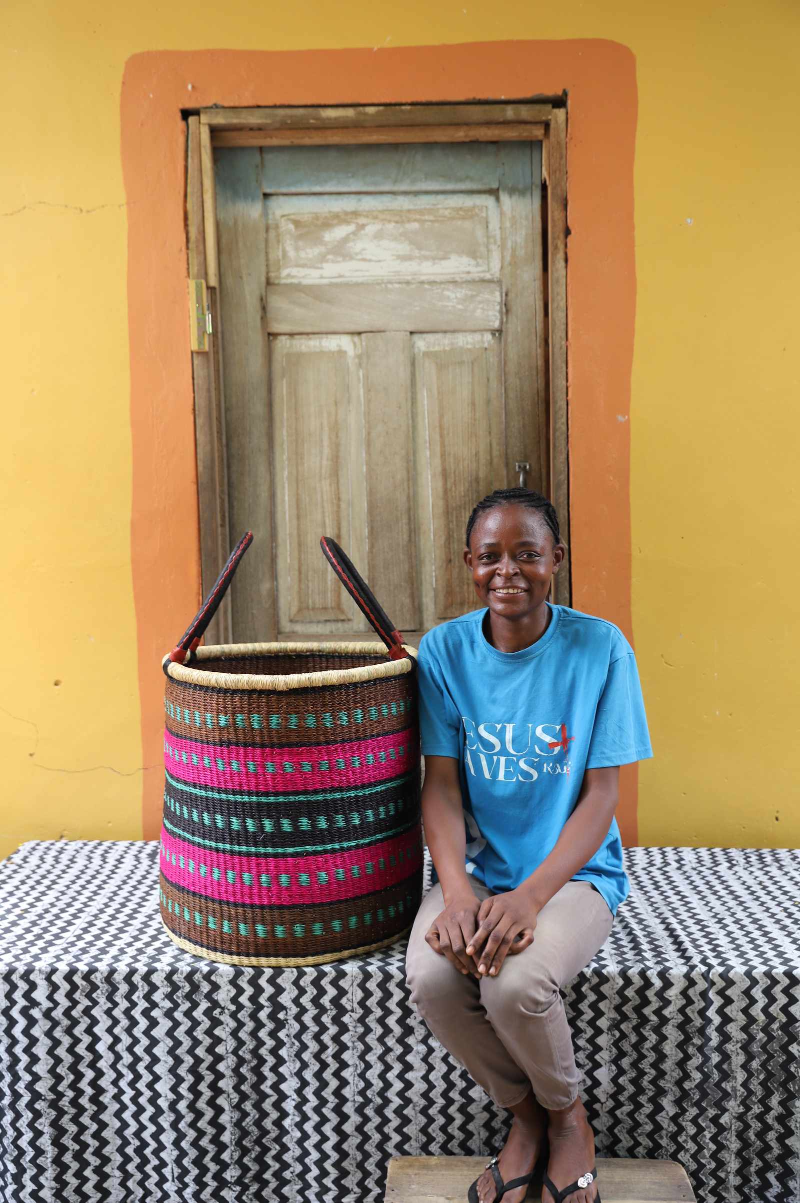 Large handwoven Bolga laundry basket in natural with pink and dark bands, beside a seated woman in blue.