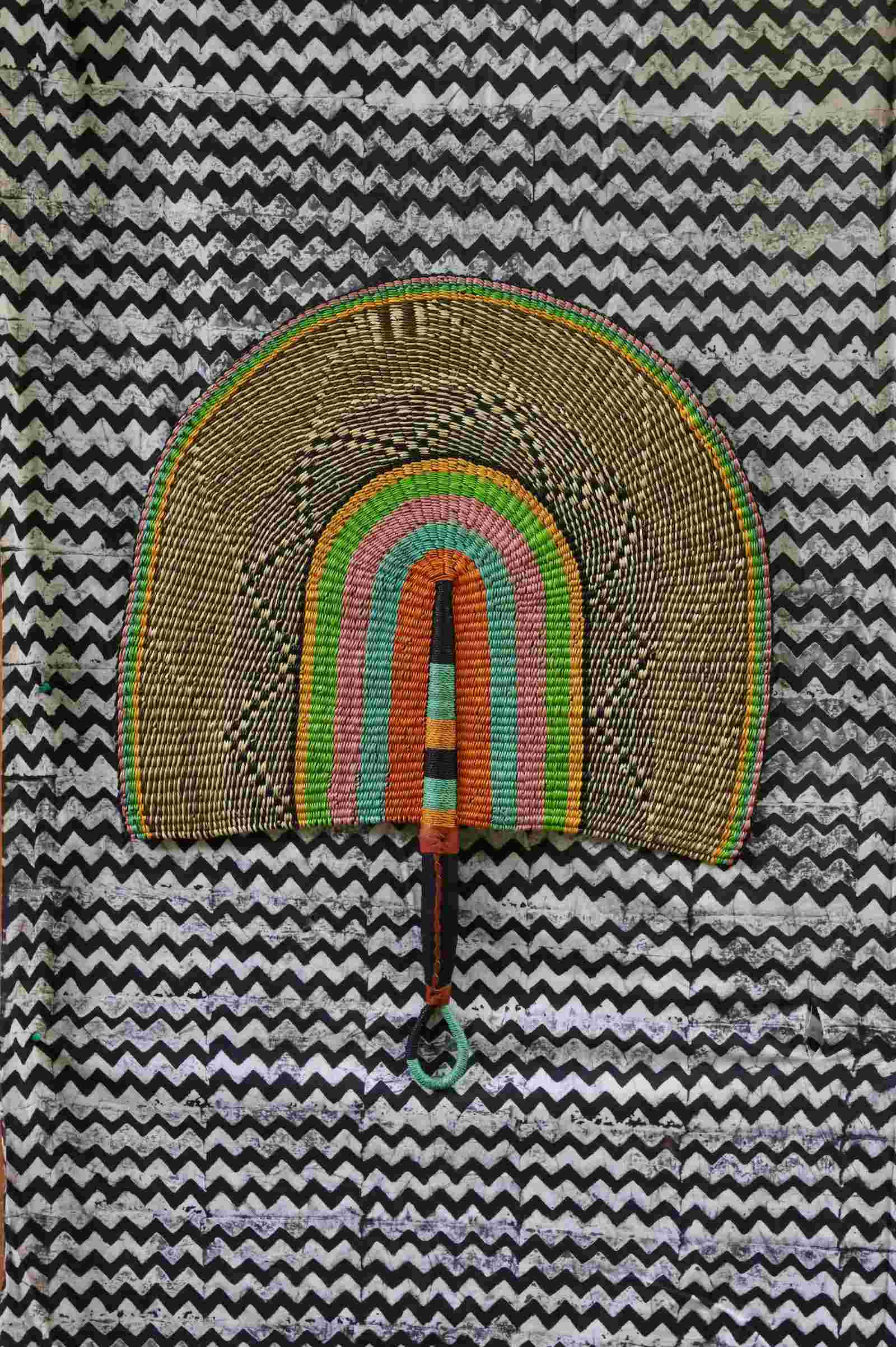 Naba Penga handwoven wall fan with natural straw outer arch and inner bands of pink, teal and orange, on a chevron backdrop.