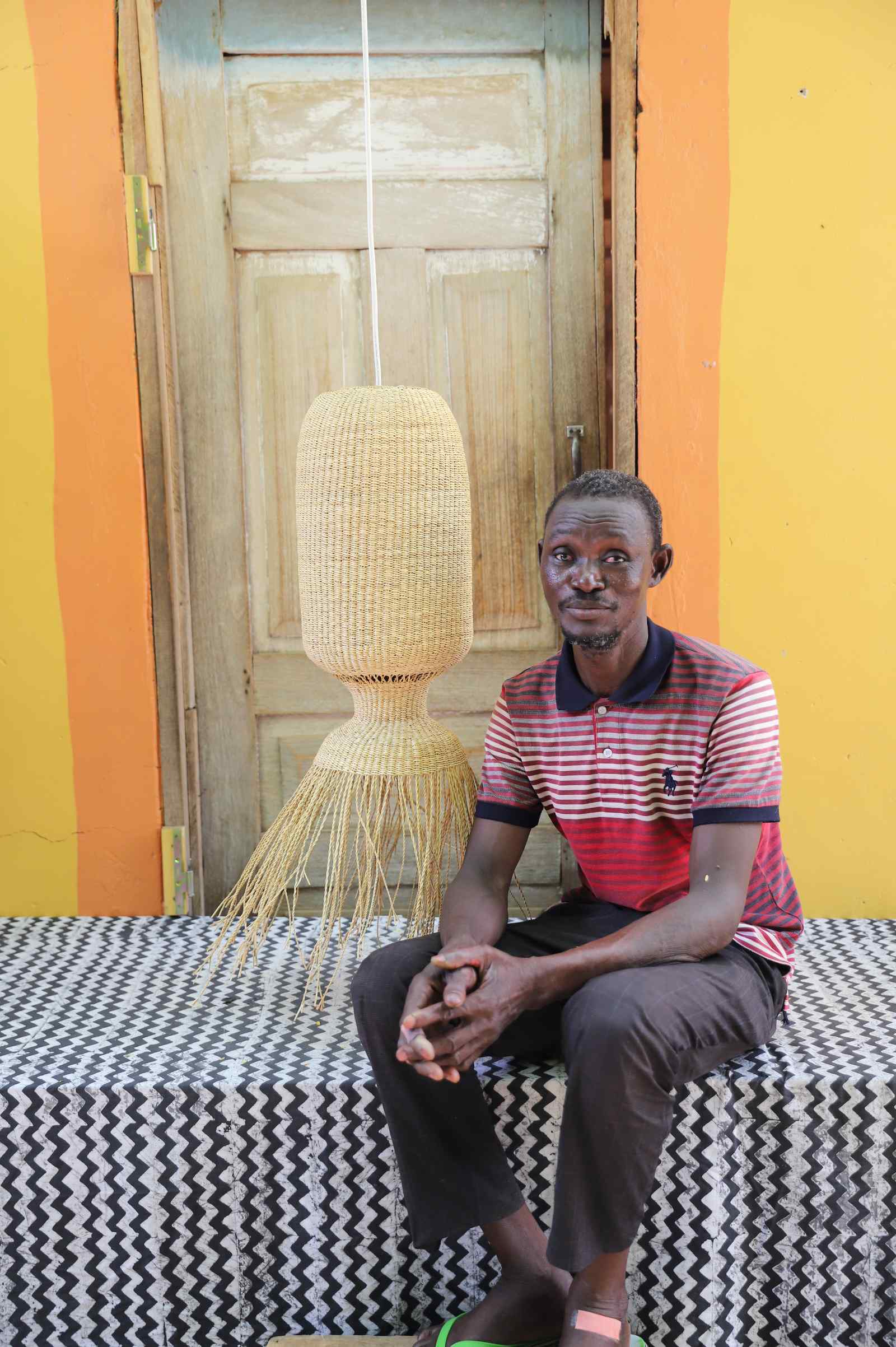 Man seated on a woven bench beside the Abyssal lampshade in a weathered doorway.