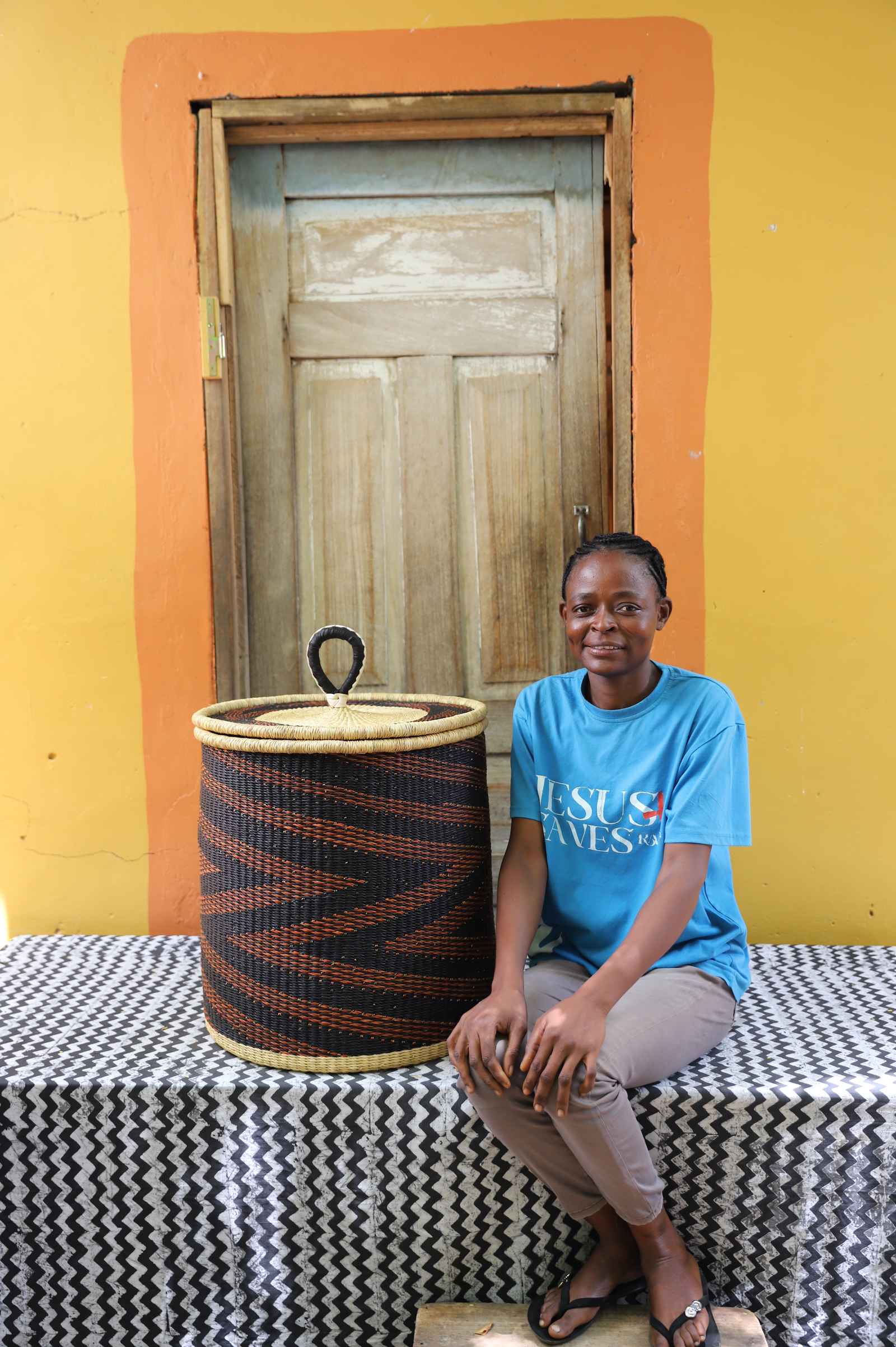 Handwoven Bolga laundry basket in dark navy with rust-orange waves, beside a seated woman in blue.