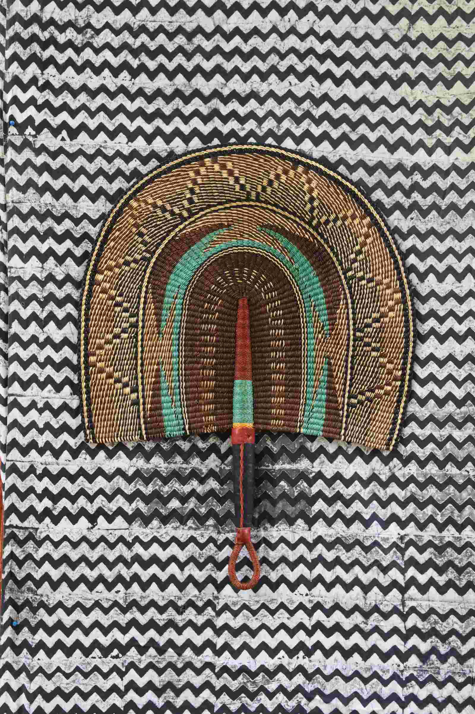 Naba Penga wall fan in natural straw with a teal inner arch and dark patterned border, laid on a chevron cloth.