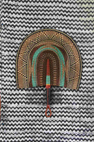 Naba Penga wall fan in natural straw with a teal inner arch and dark patterned border, laid on a chevron cloth.