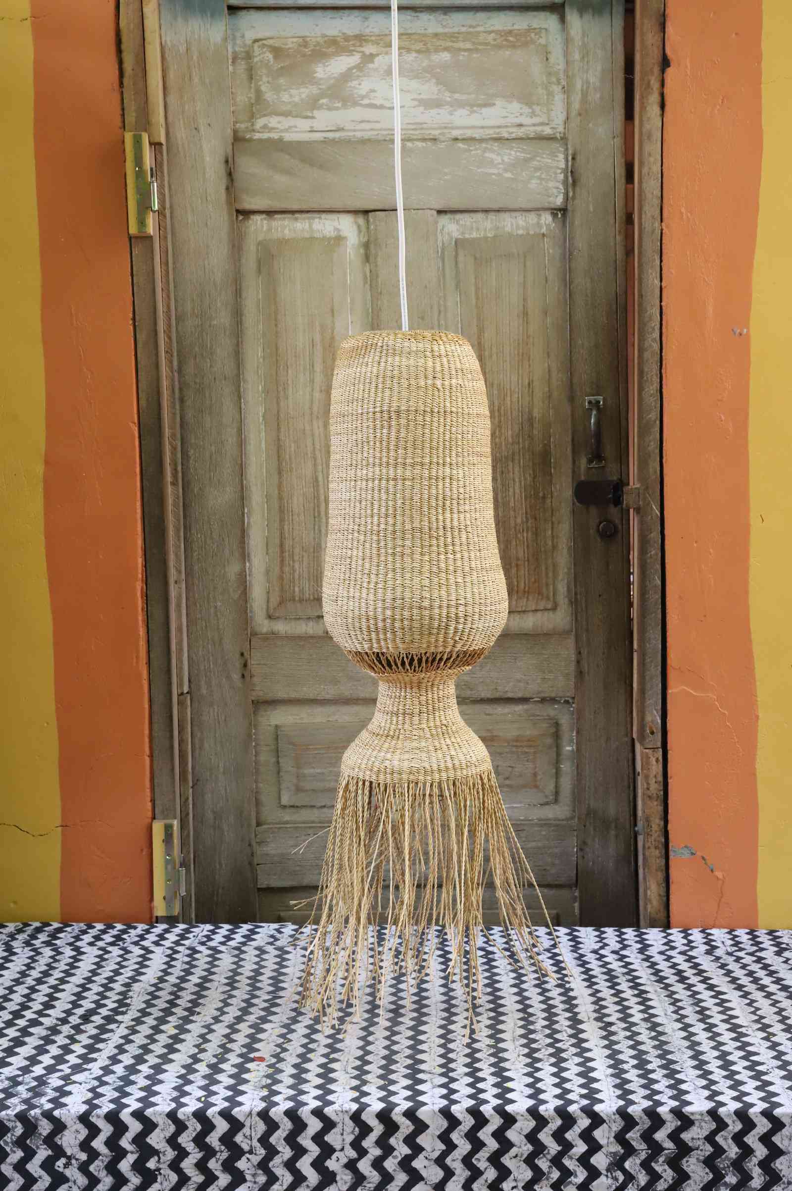Elephant grass Abyssal pendant lampshade with a bulbed waist and neat fringe, hung against a wooden door.