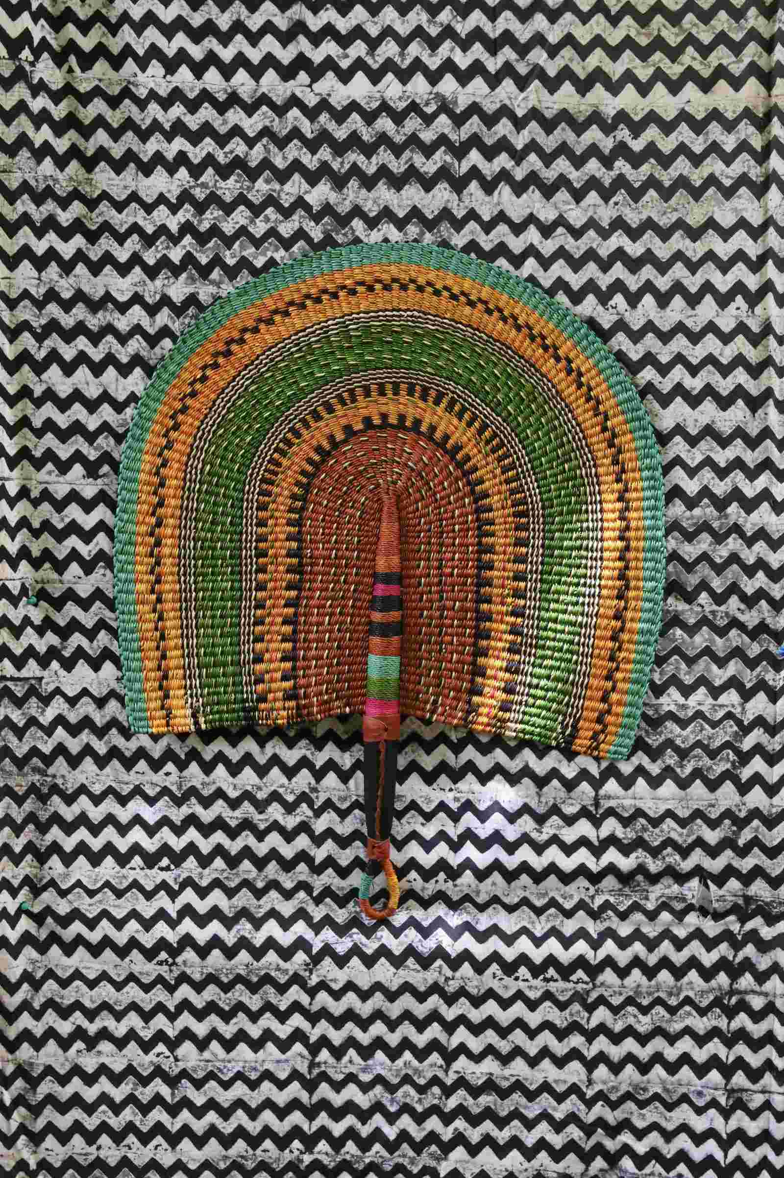 Naba Penga handwoven wall fan with arch bands in green, amber and terracotta, on a black-and-white chevron backdrop.