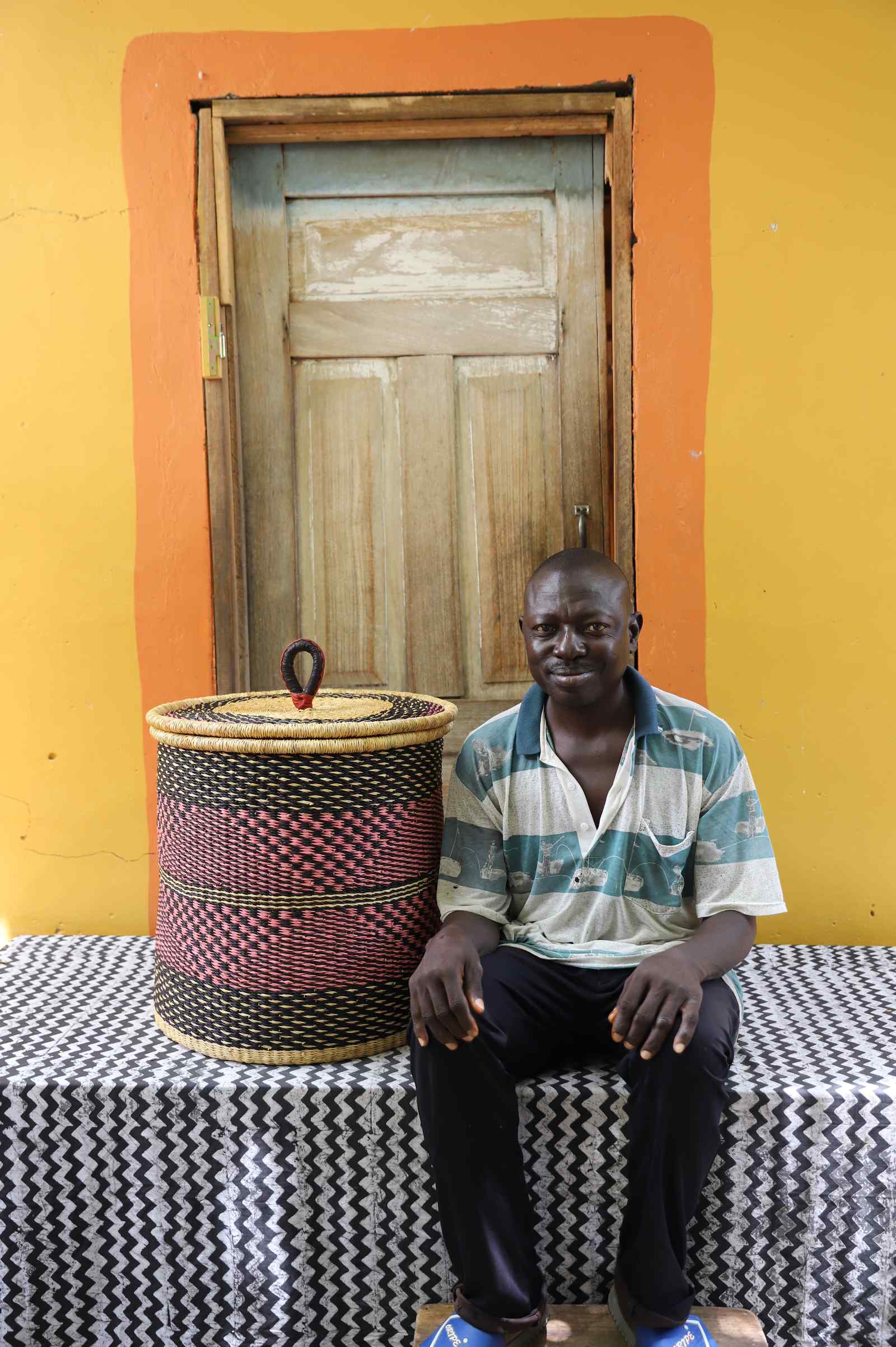 Handwoven Bolga laundry basket in natural with red-pink checkered band, beside a seated man in stripes.