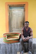 Large handwoven Bolga pet bed in natural straw with green, red and orange bands, beside a seated man.