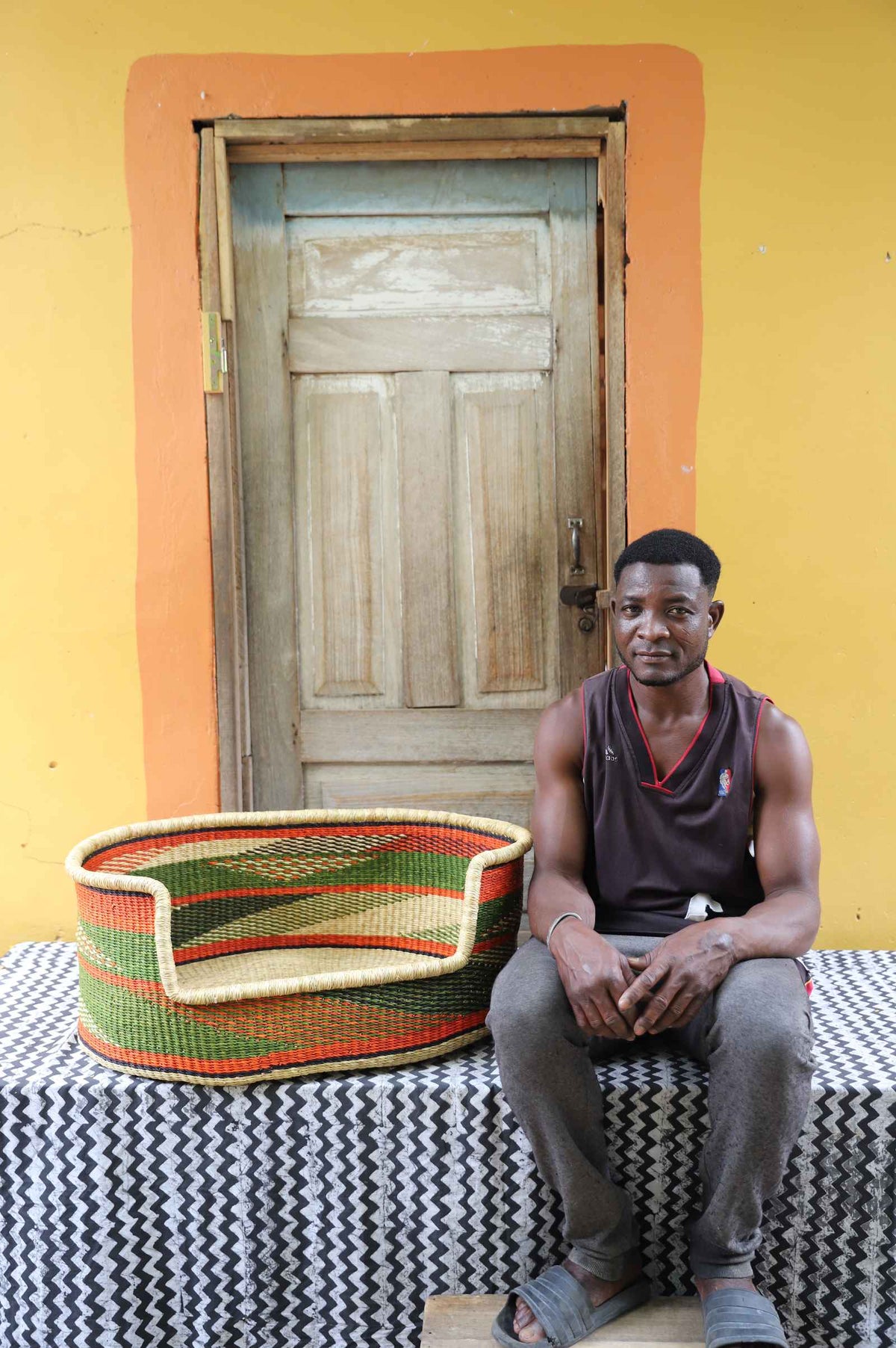 Large handwoven Bolga pet bed in natural straw with green, red and orange bands, beside a seated man.