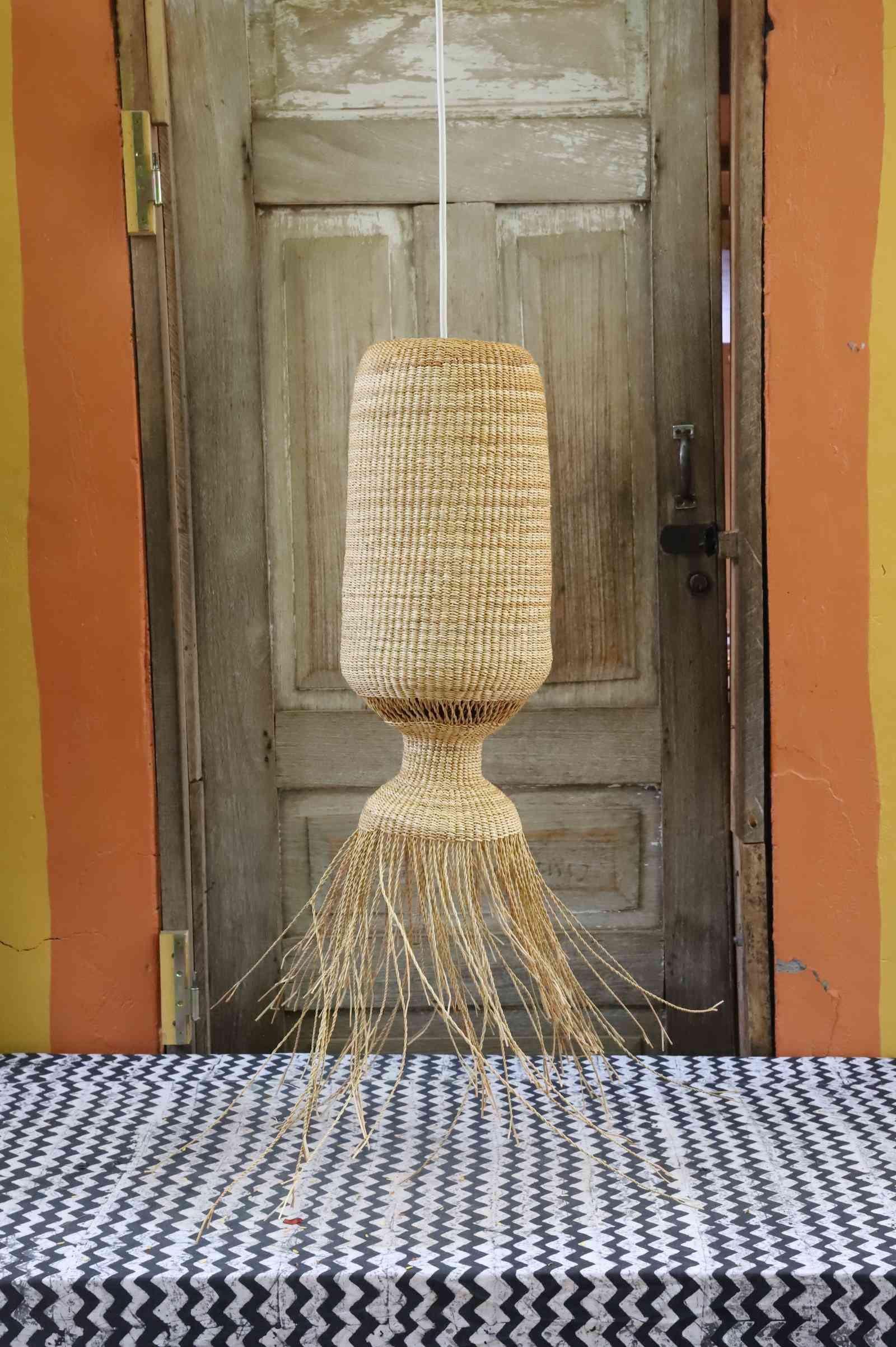 Abyssal pendant lampshade in woven elephant grass with a fringed base, suspended before a weathered wooden door.