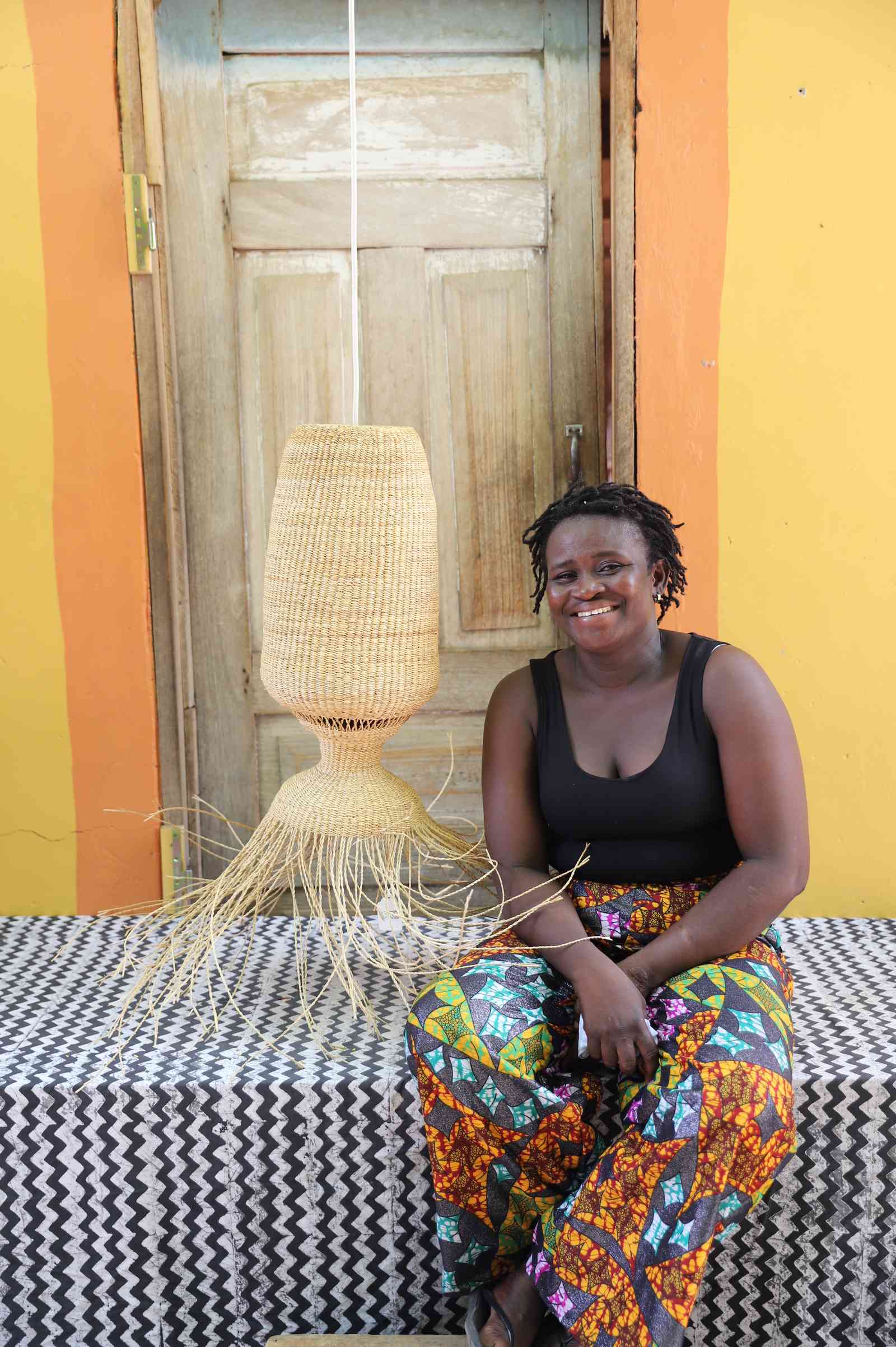 Woman seated on a woven bench beside the Abyssal lampshade in a weathered doorway.