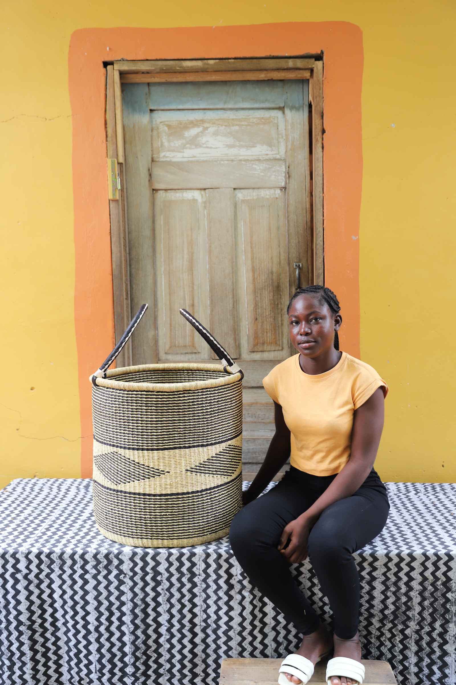 Large handwoven Bolga laundry basket in natural with dark diamond motif, beside a seated woman in yellow.