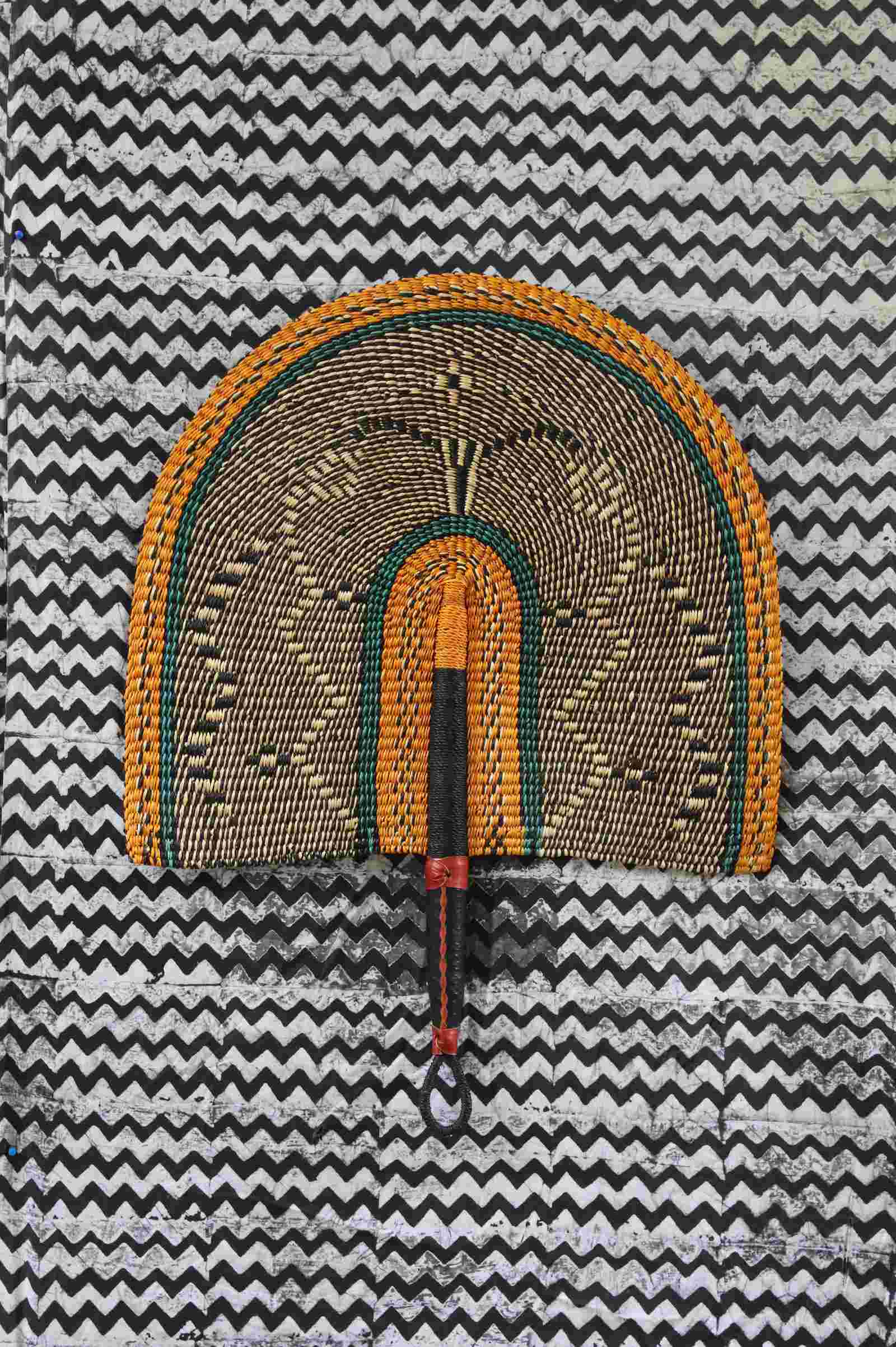 Naba Penga wall fan in natural straw with a mustard border and green trim, laid on a chevron cloth.