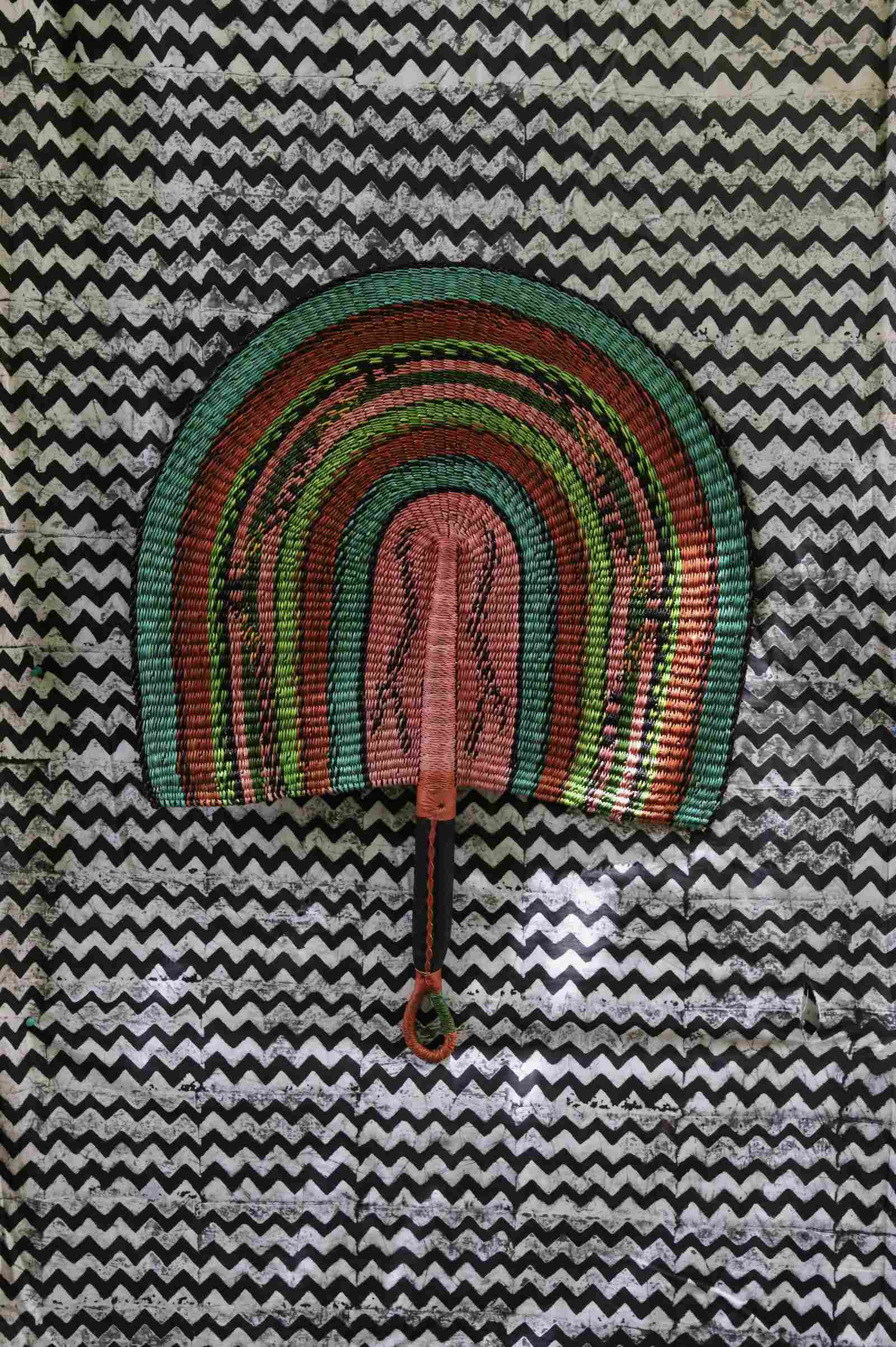 Naba Penga handwoven wall fan in teal, terracotta and dusty pink banded stripes, on a chevron-print backdrop.