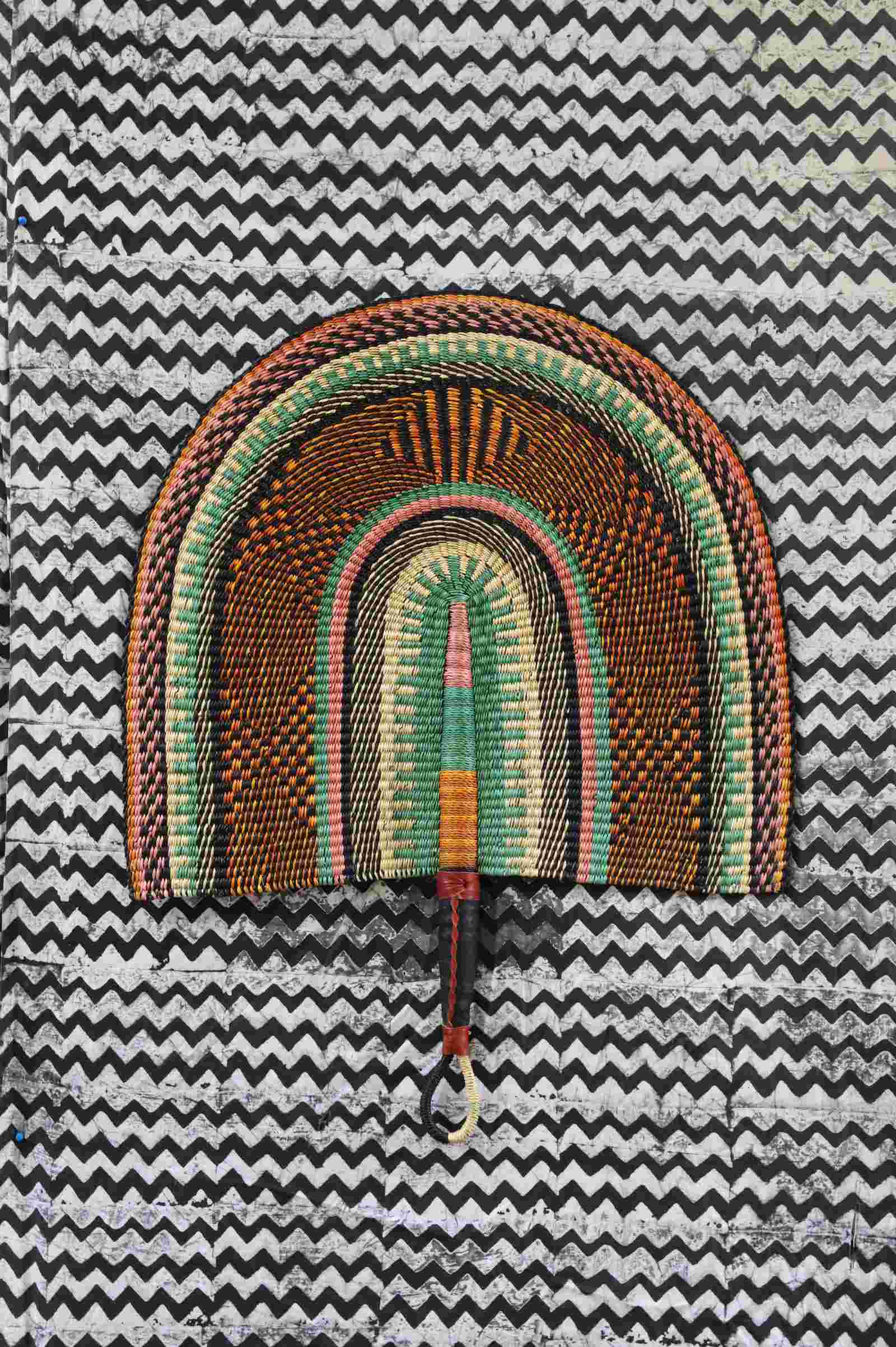 Naba Penga wall fan in natural straw with sage green and rust banded arches, laid on a chevron cloth.