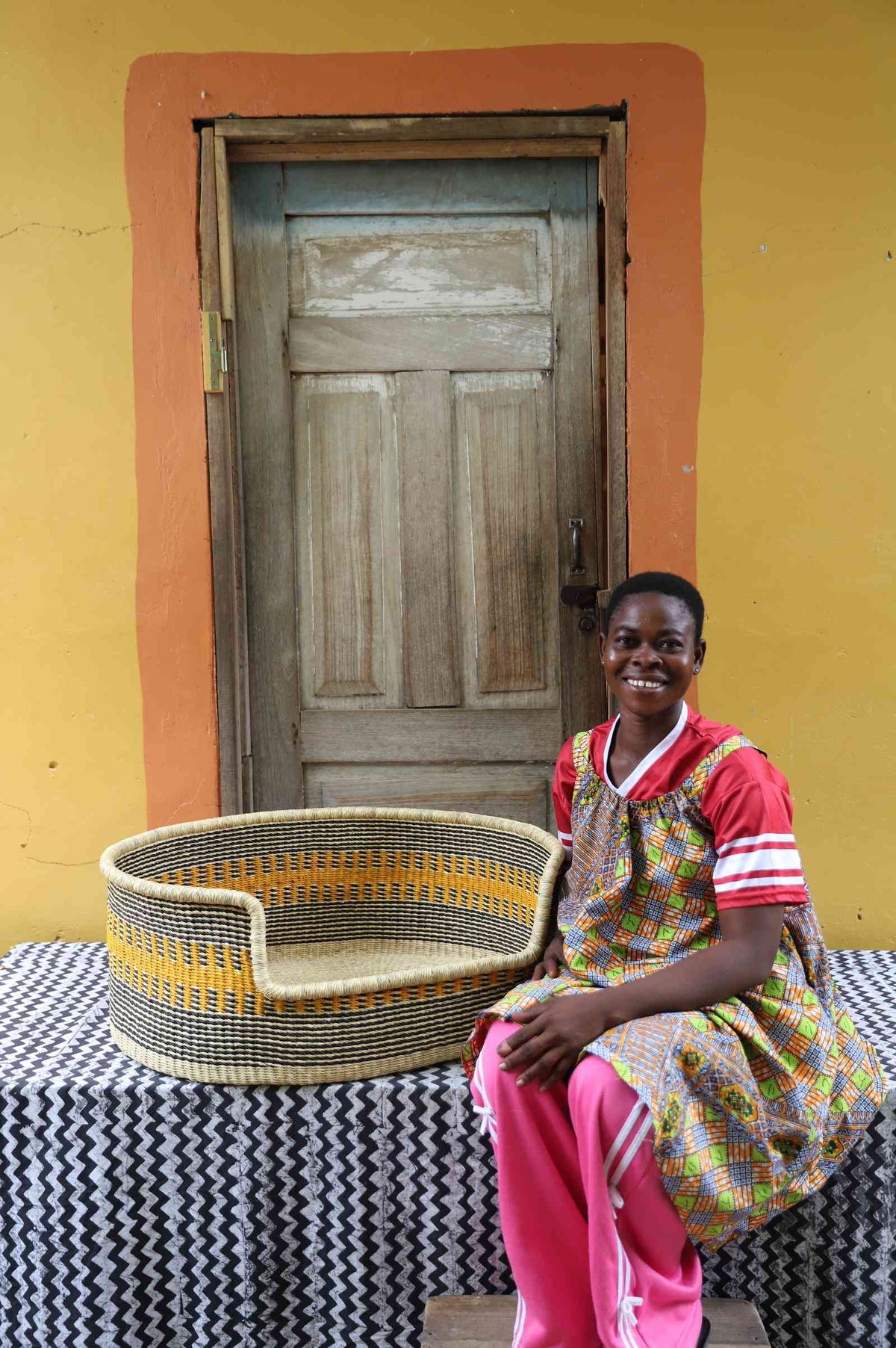 Large handwoven Bolga pet bed in natural straw with orange and yellow bands, beside a seated woman in red.
