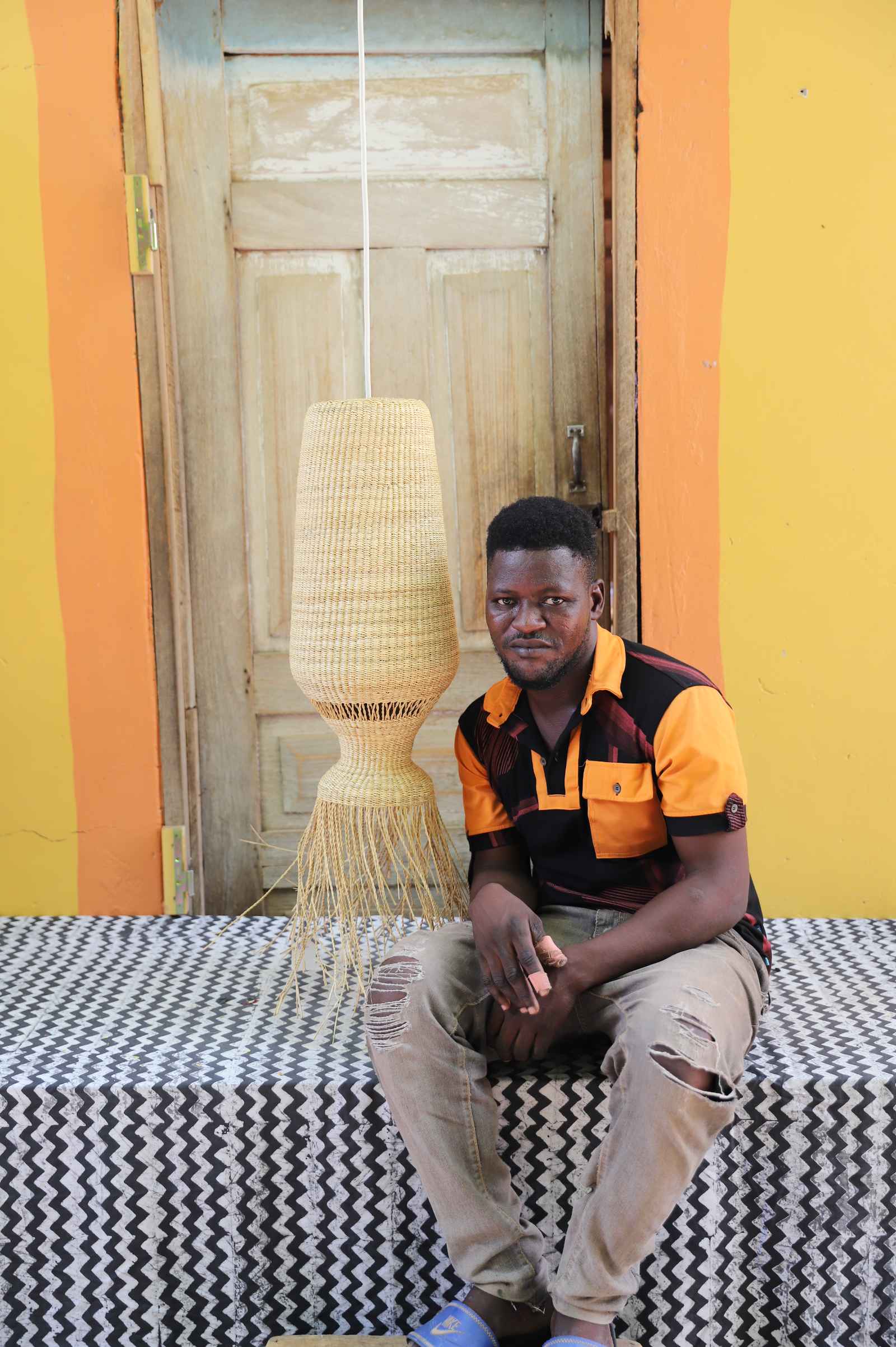 Man seated on a woven stool beneath the Abyssal lampshade in a weathered wooden doorway.