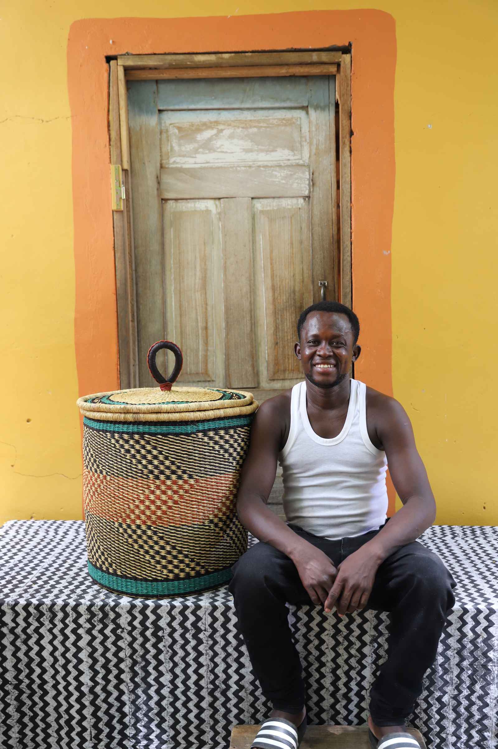 Handwoven Bolga laundry basket in natural with orange and dark bands, beside a seated man in a white vest.