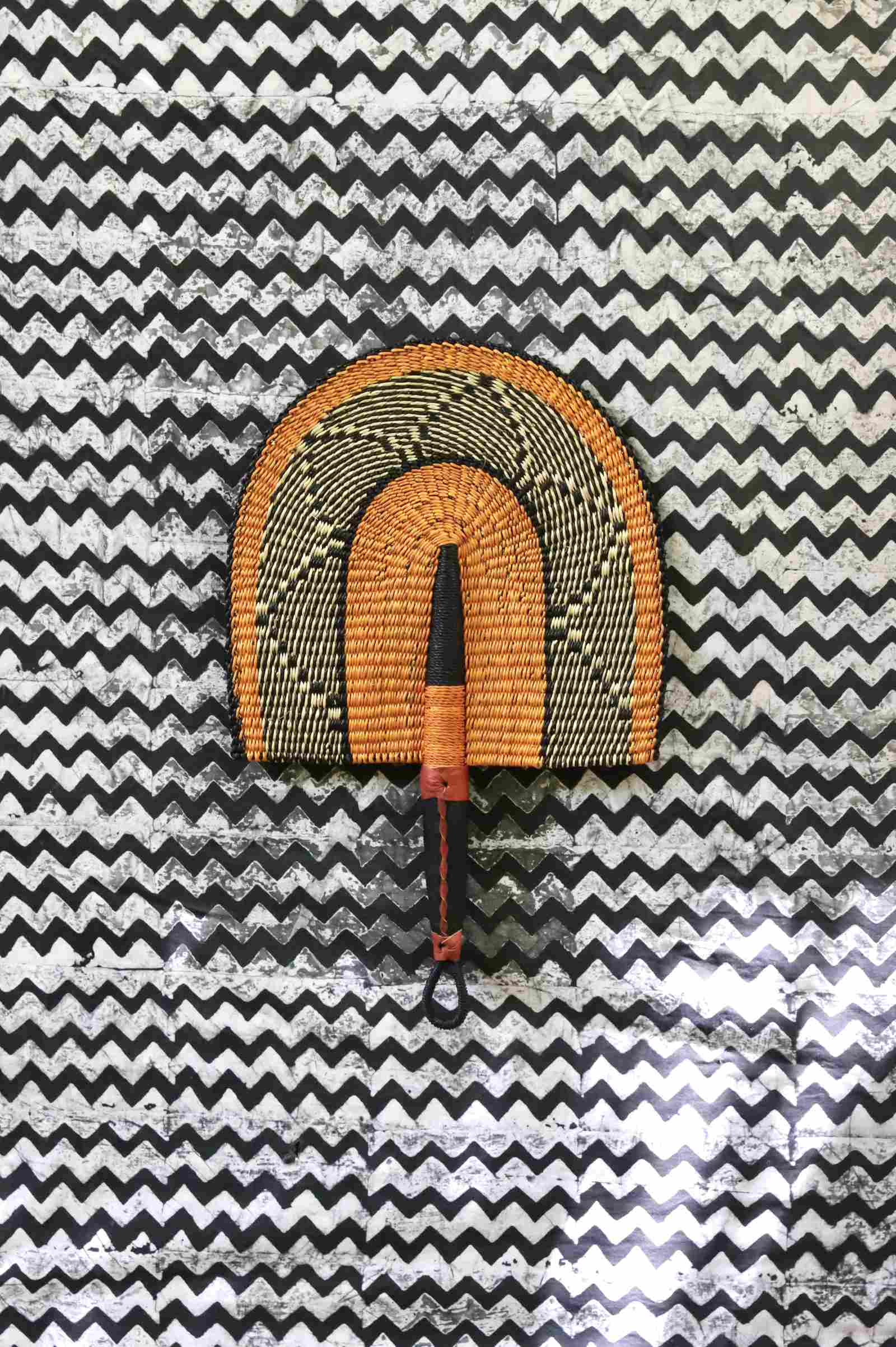 Handwoven Bolga Fan in natural straw with black geometric outer arch, on a black-and-white chevron fabric backdrop.