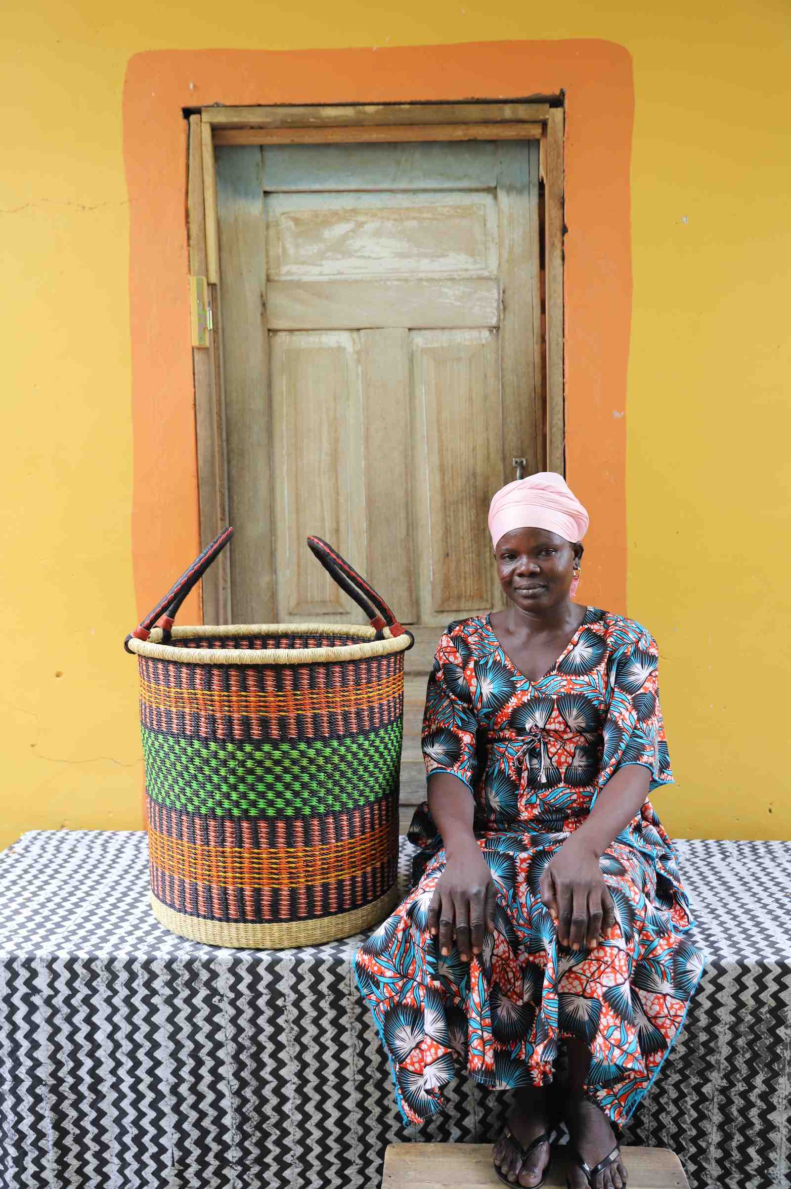 Large handwoven Bolga laundry basket in dark straw with green and orange bands, beside a seated woman in floral.