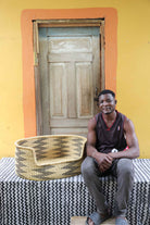 Large handwoven Bolga pet bed in natural straw with black arrows, beside a seated man in a dark vest.