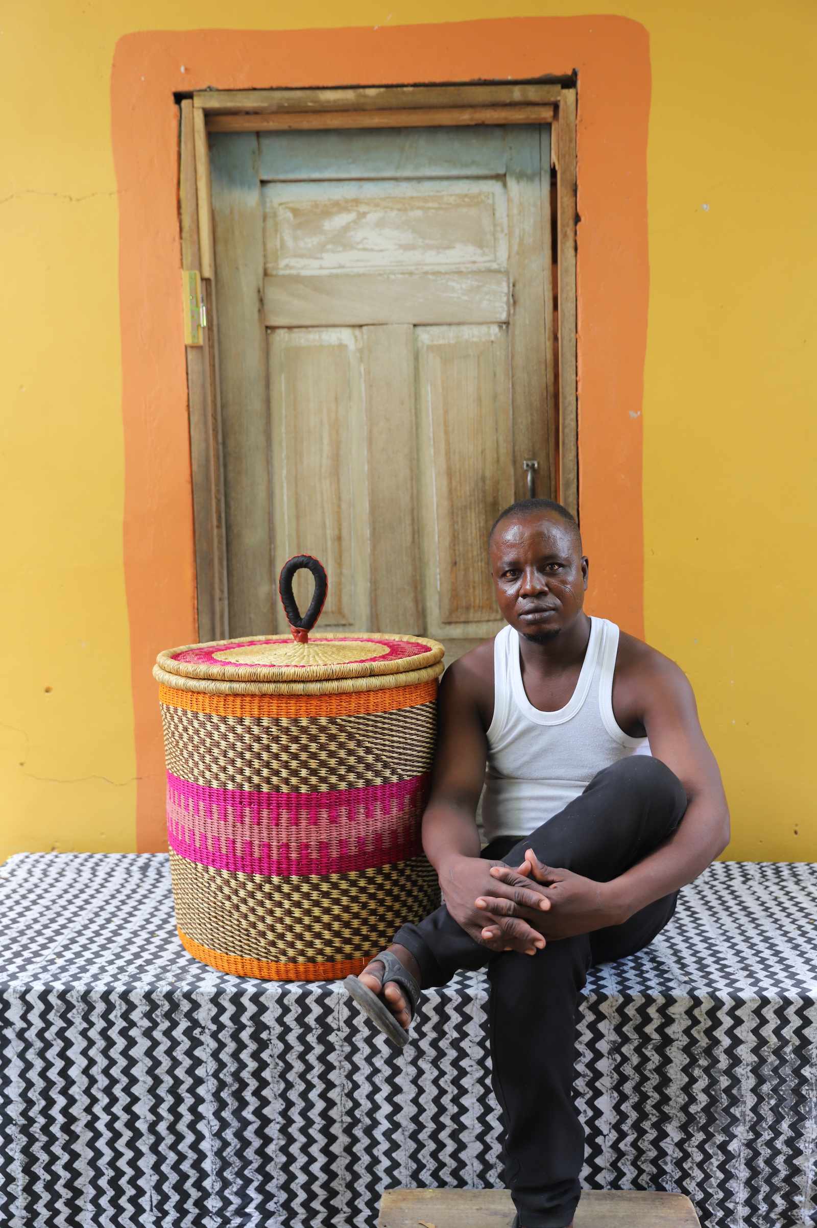 Handwoven Bolga laundry basket with pink and orange central bands, beside a seated man in a white vest.