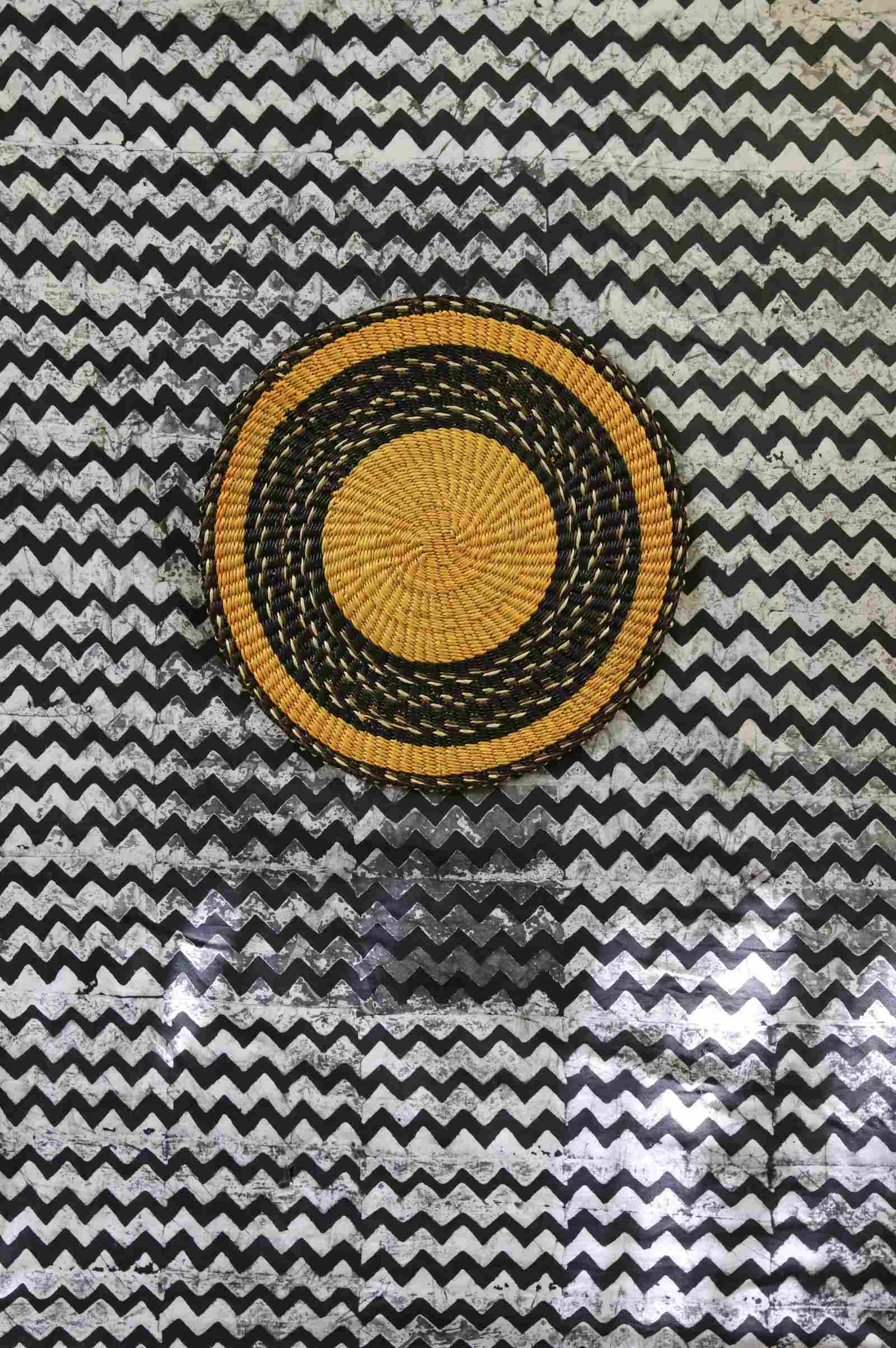 Handwoven Bolga placemat in natural straw with bold black border ring, on a black-and-white chevron fabric backdrop.