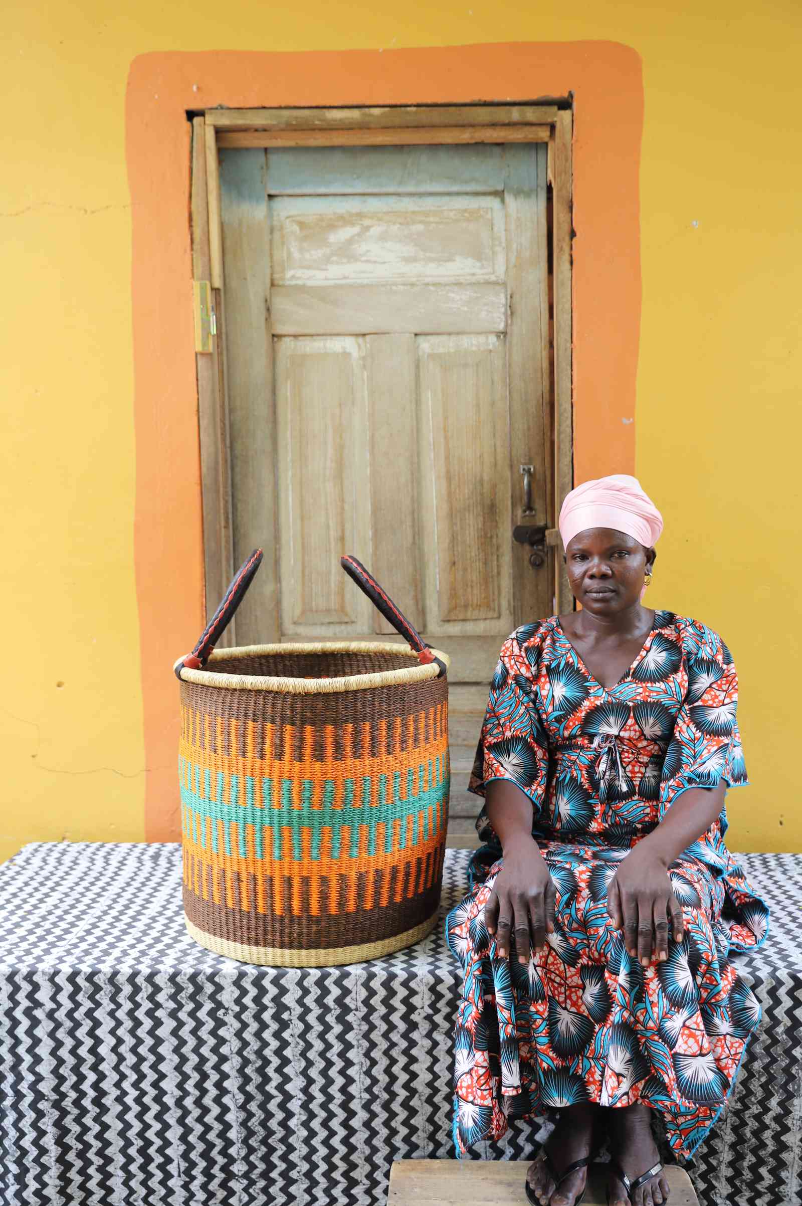 Large handwoven Bolga laundry basket in brown and orange bands, beside a seated woman in a printed dress.