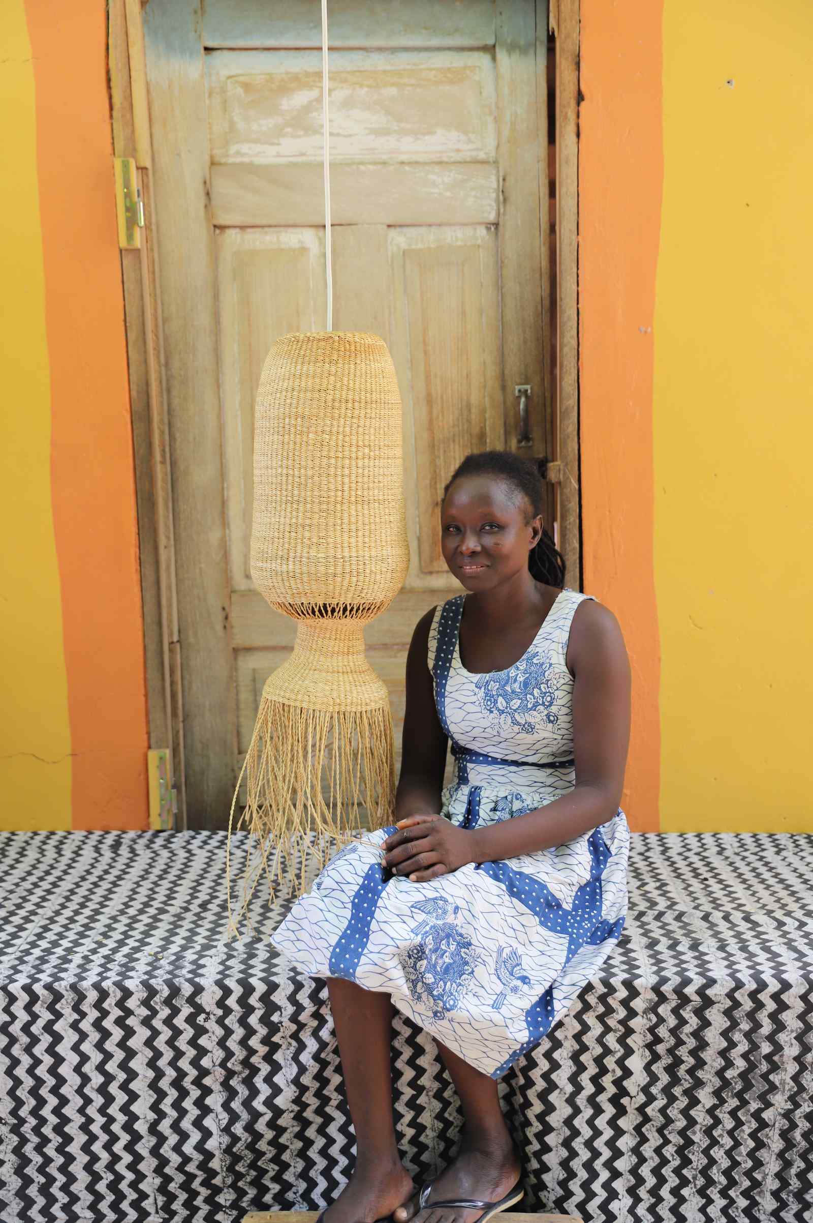 Woman in a patterned dress seated beside the Abyssal lampshade in a weathered doorway.
