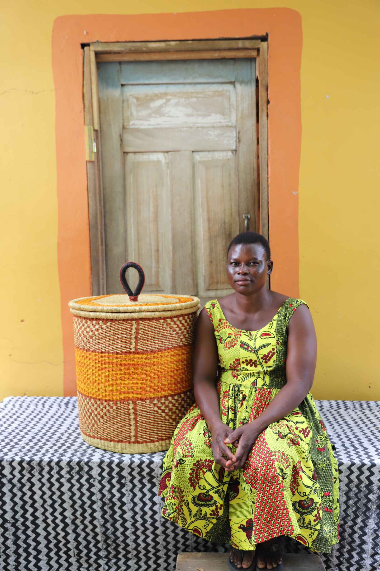 Handwoven Bolga laundry basket with orange central band, beside a seated woman in green and yellow.