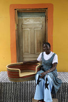 Medium handwoven Bolga pet bed in natural straw with red bands, beside a seated woman in a printed dress.