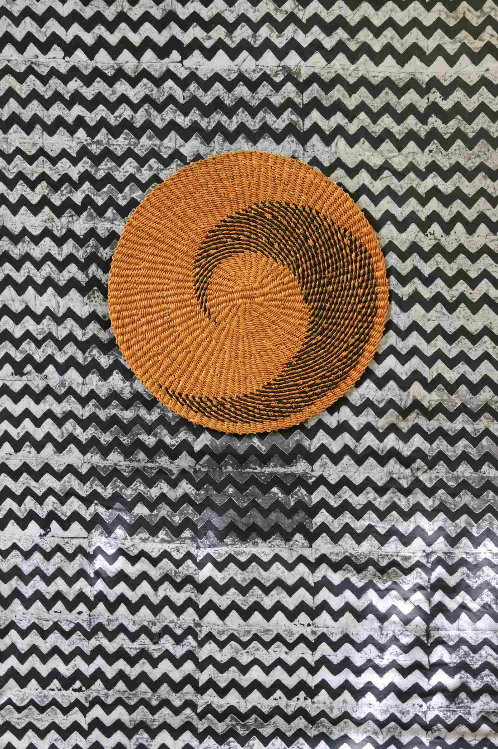 Handwoven Bolga placemat in natural straw with black spiral pattern, on a black-and-white chevron fabric backdrop.
