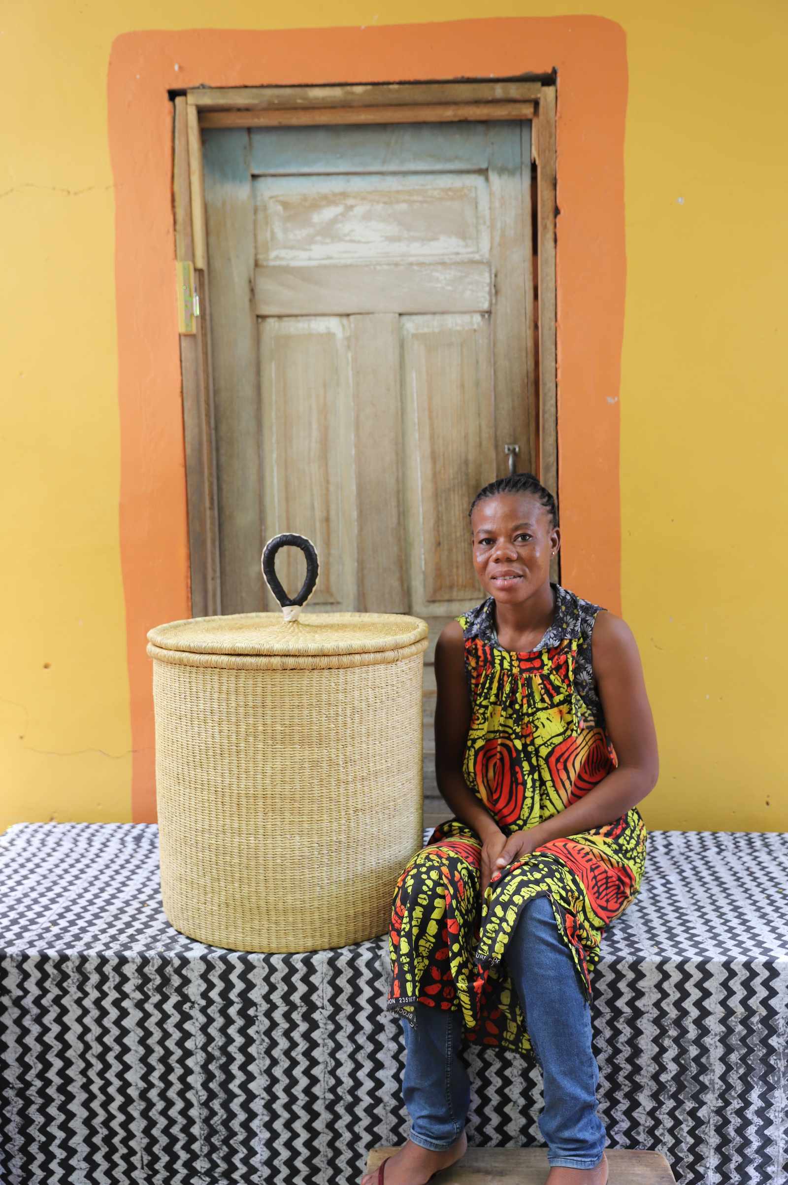 Handwoven Bolga laundry basket in plain natural straw, beside a seated woman in a printed dress.
