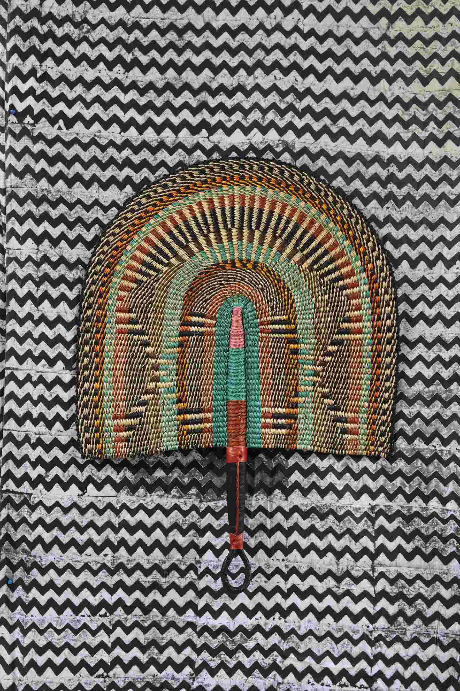 Naba Penga wall fan in natural straw with rust, green and teal concentric arches, laid on a chevron cloth.